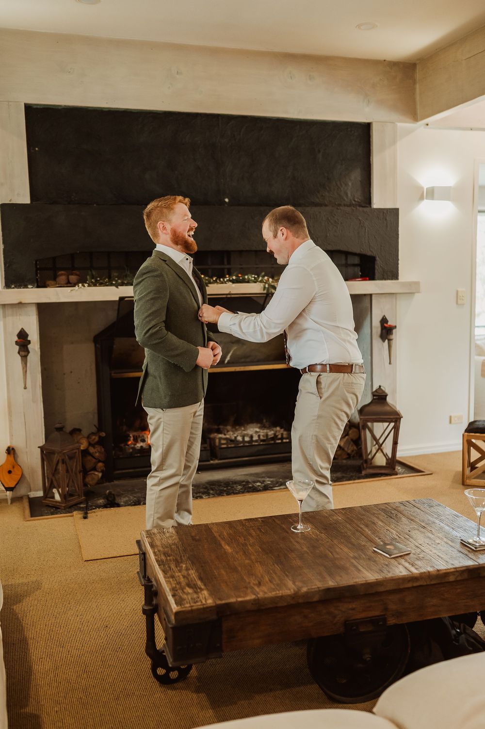 Two men in formal attire laugh together indoors by a fireplace, one adjusting the other's jacket lapel.