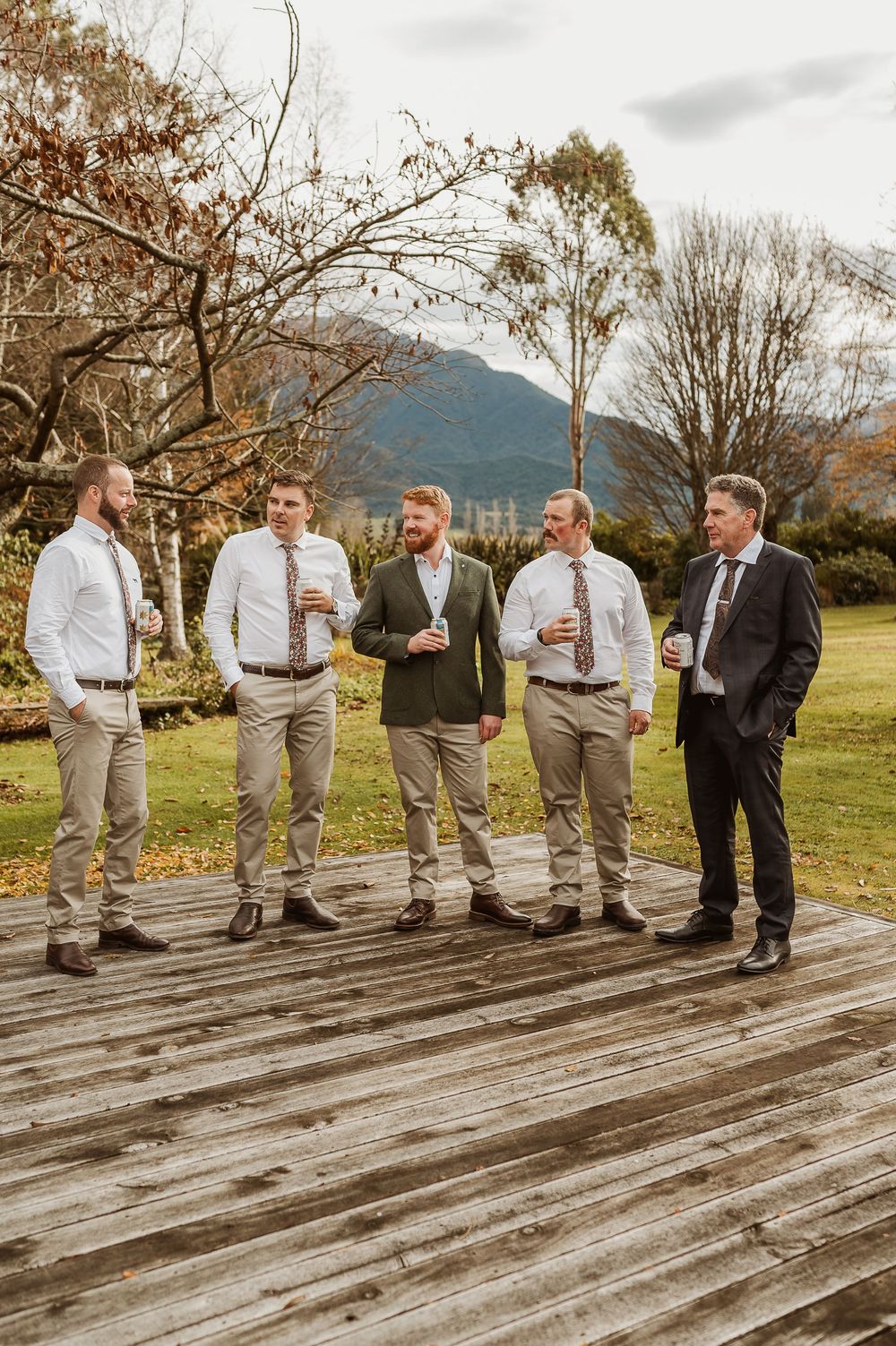 Groom and groomsmen in khaki pants and ties toasting drinks on wooden deck with mountain backdrop.