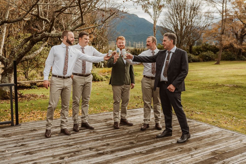 Five groomsmen raising glasses in a celebratory toast outdoors on a wooden deck with autumn trees.