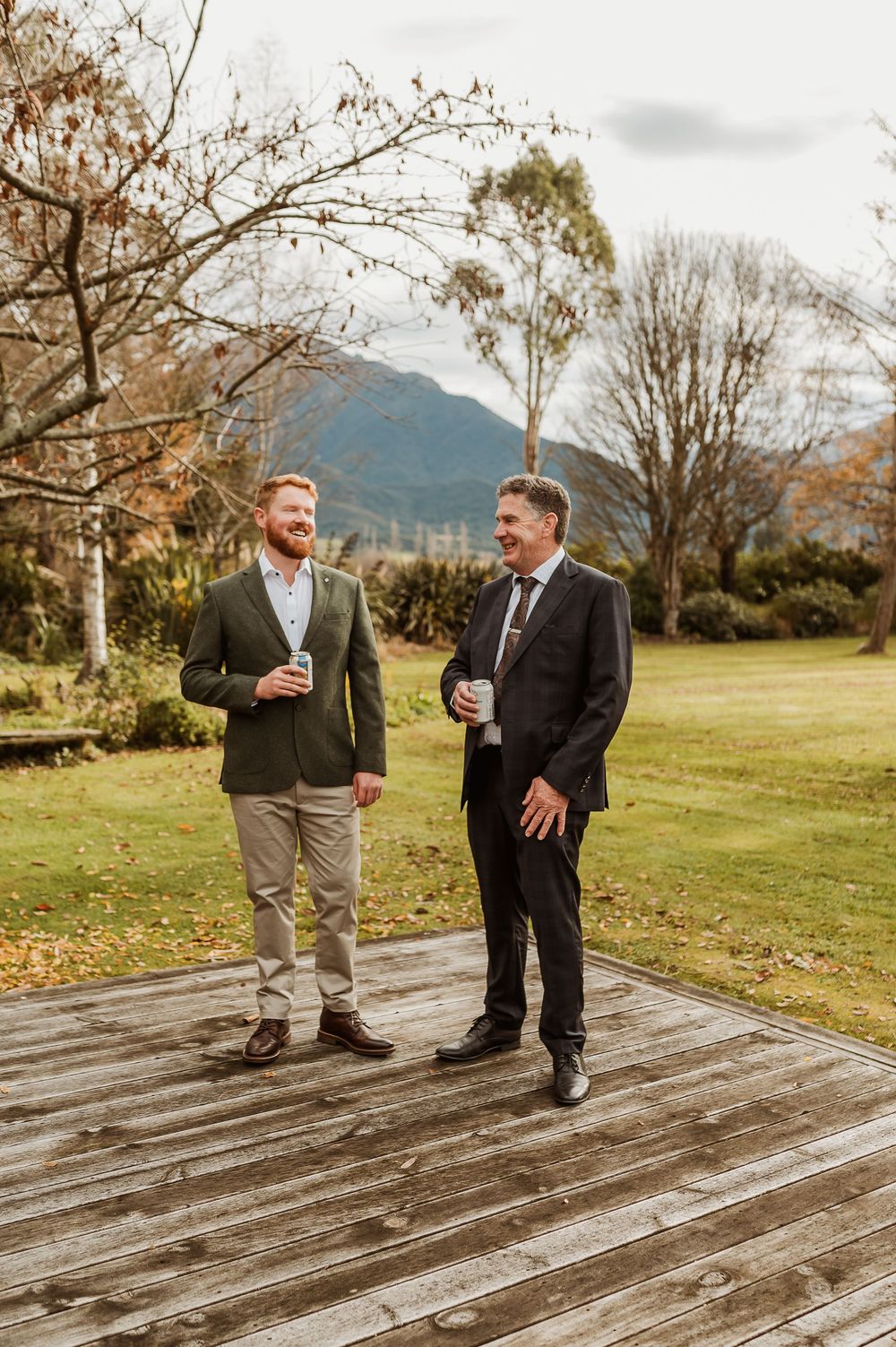 Two well-dressed men smile and chat on a wooden deck surrounded by autumn trees and mountain views.