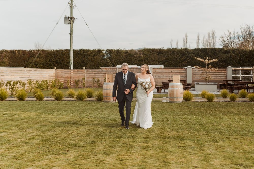 Bride and groom walking across green lawn at outdoor wedding venue with wooden barrels and hedges.