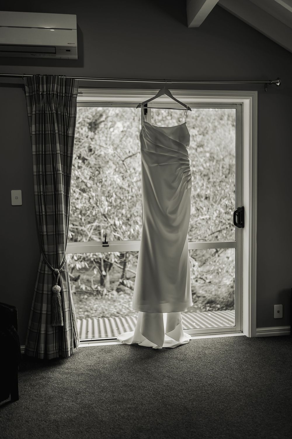 Elegant white wedding dress hanging on a hanger in front of a window in a cozy room.
