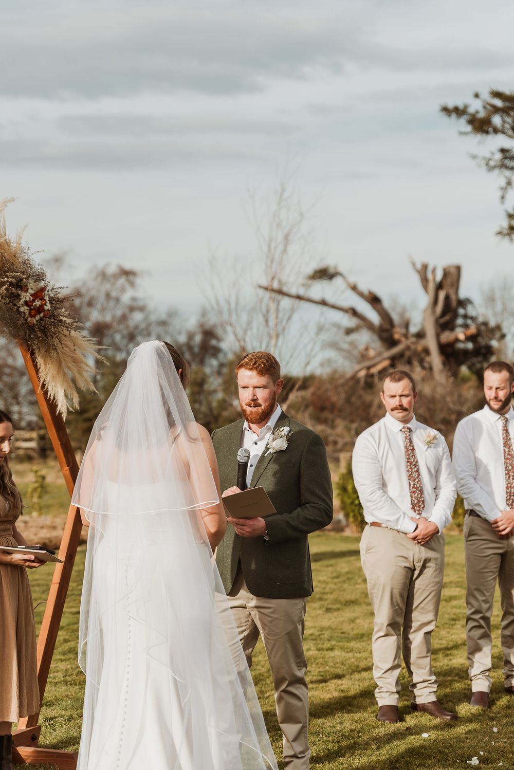 Outdoor wedding ceremony with bride in white veil, groom in green suit holding book, and groomsmen standing on green lawn.