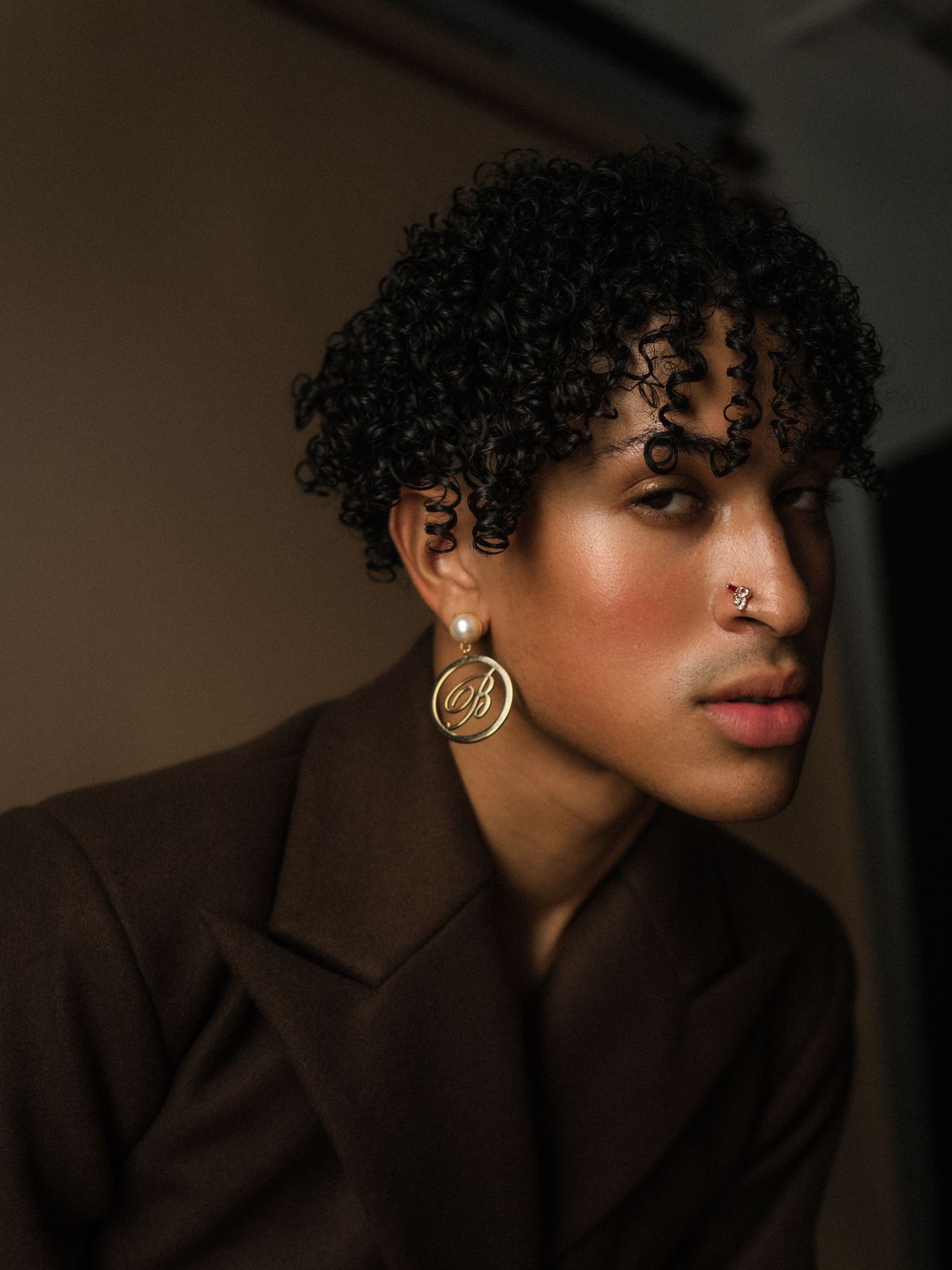 Person with curly hair wearing gold monogram earrings and a brown blazer, with a nose ring.