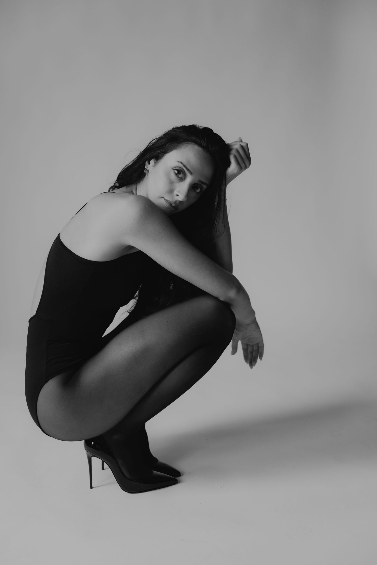 Black and white studio photo of a woman in a black bodysuit and heels, crouching gracefully on a light background.