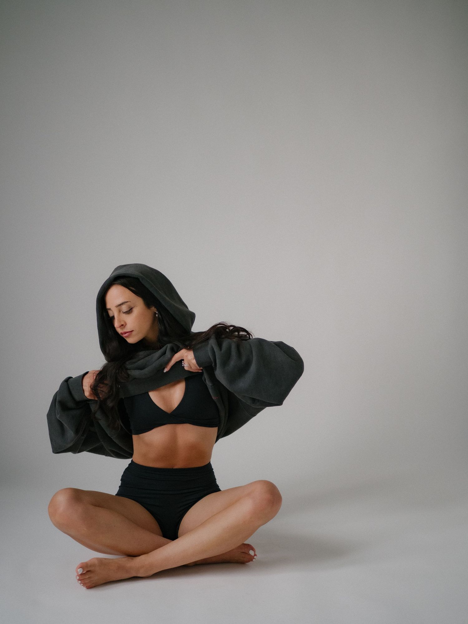 Woman in black crop top and high-waist bottoms sits cross-legged wearing oversized dark hoodie in studio setting.