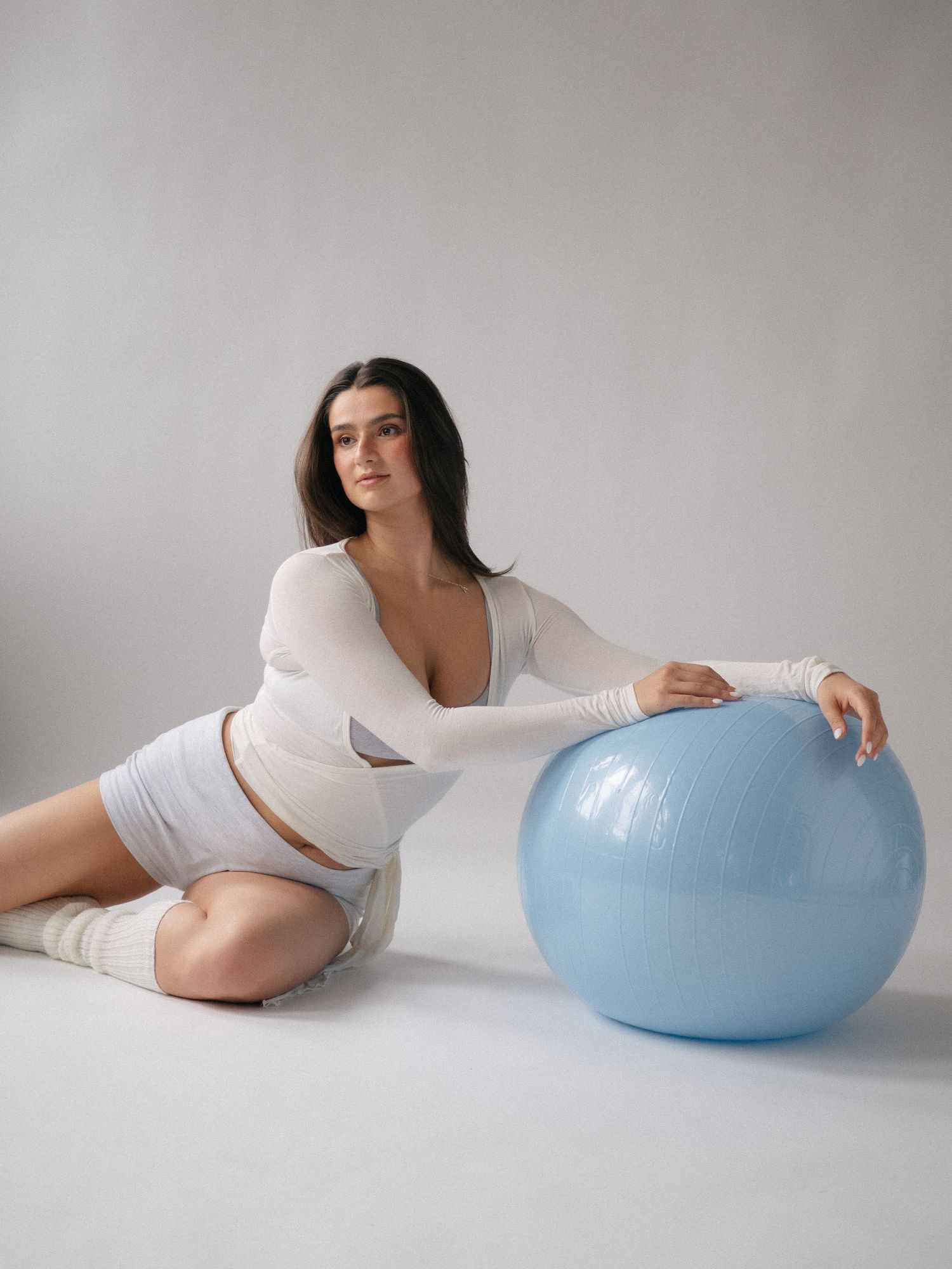 Woman in white outfit posing with a light blue exercise ball on a gray studio background.