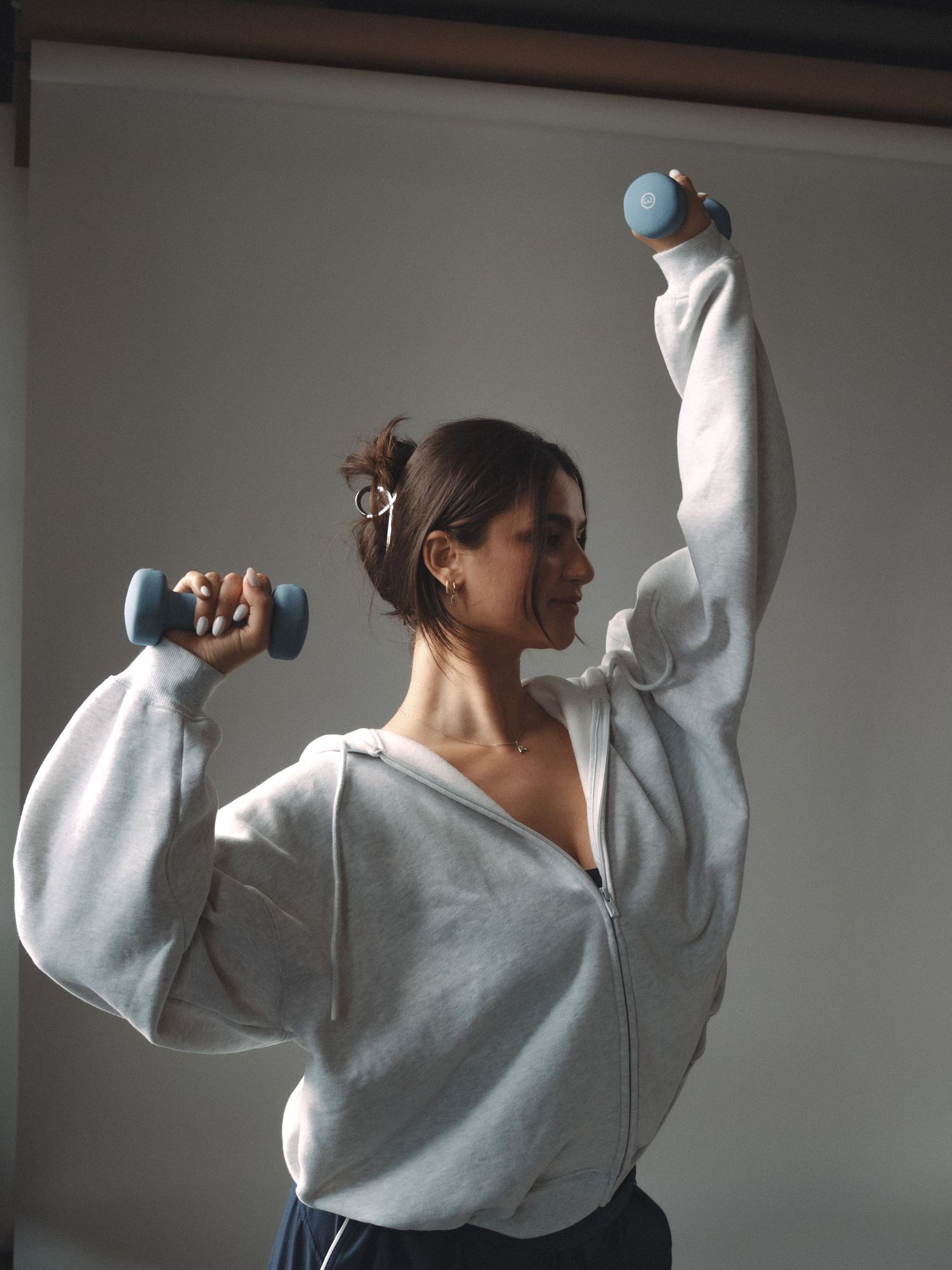 Woman in gray zip-up hoodie doing dumbbell exercises, lifting blue weights with both arms raised.