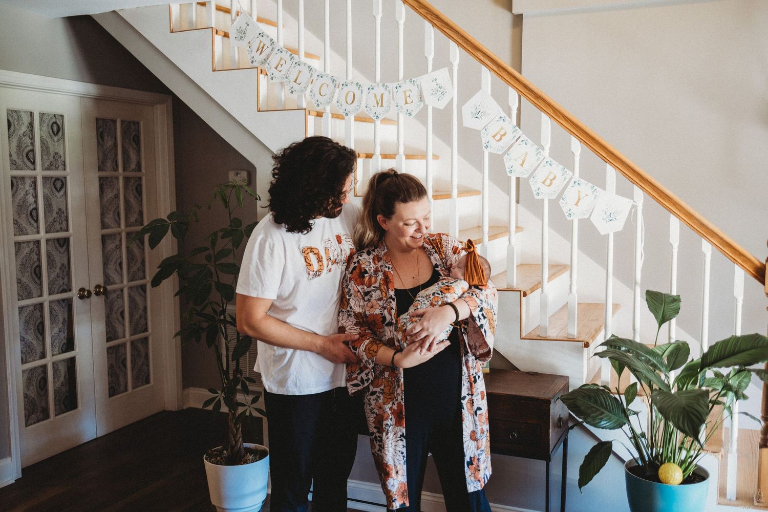 Two women admire a newborn baby indoors near a staircase decorated with welcome home bunting.