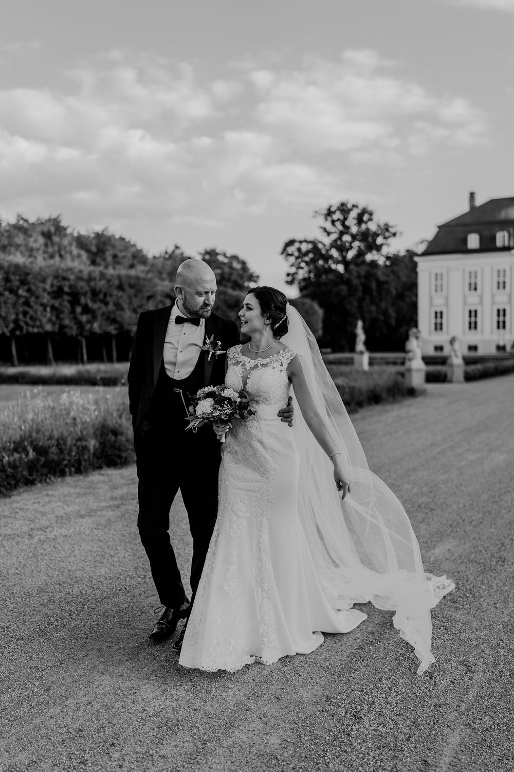 A black and white photo of a bride in a lace gown with a long veil and a groom in a tuxedo walking in a palace garden.