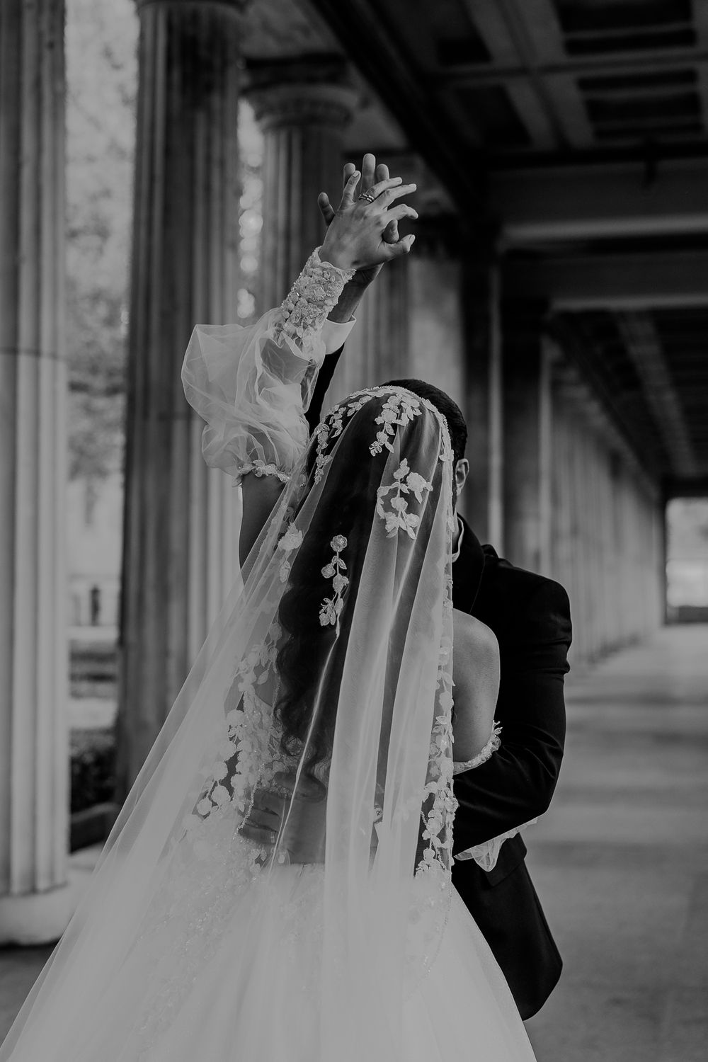 Black and white image of a bride in a lace veil raising her hand beside a groom under stone columns.