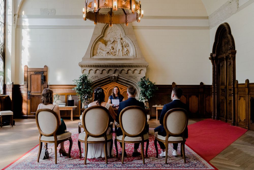 Guests and a officiant sit on cushioned chairs around a small table during an intimate civil wedding ceremony beneath a chandelier.