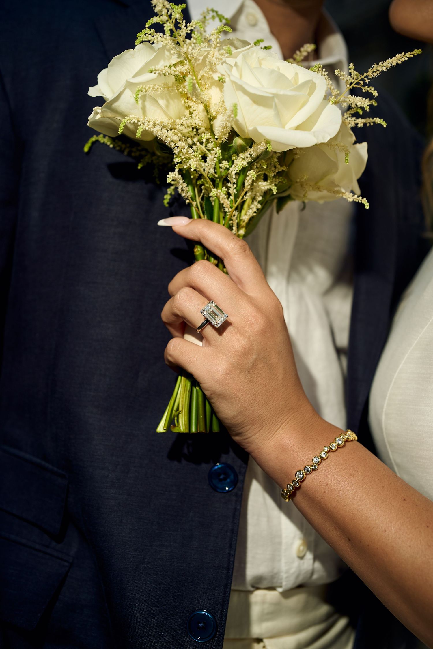 Bride holding white rose bouquet with elegant diamond ring and gold bracelet beside groom in navy suit.