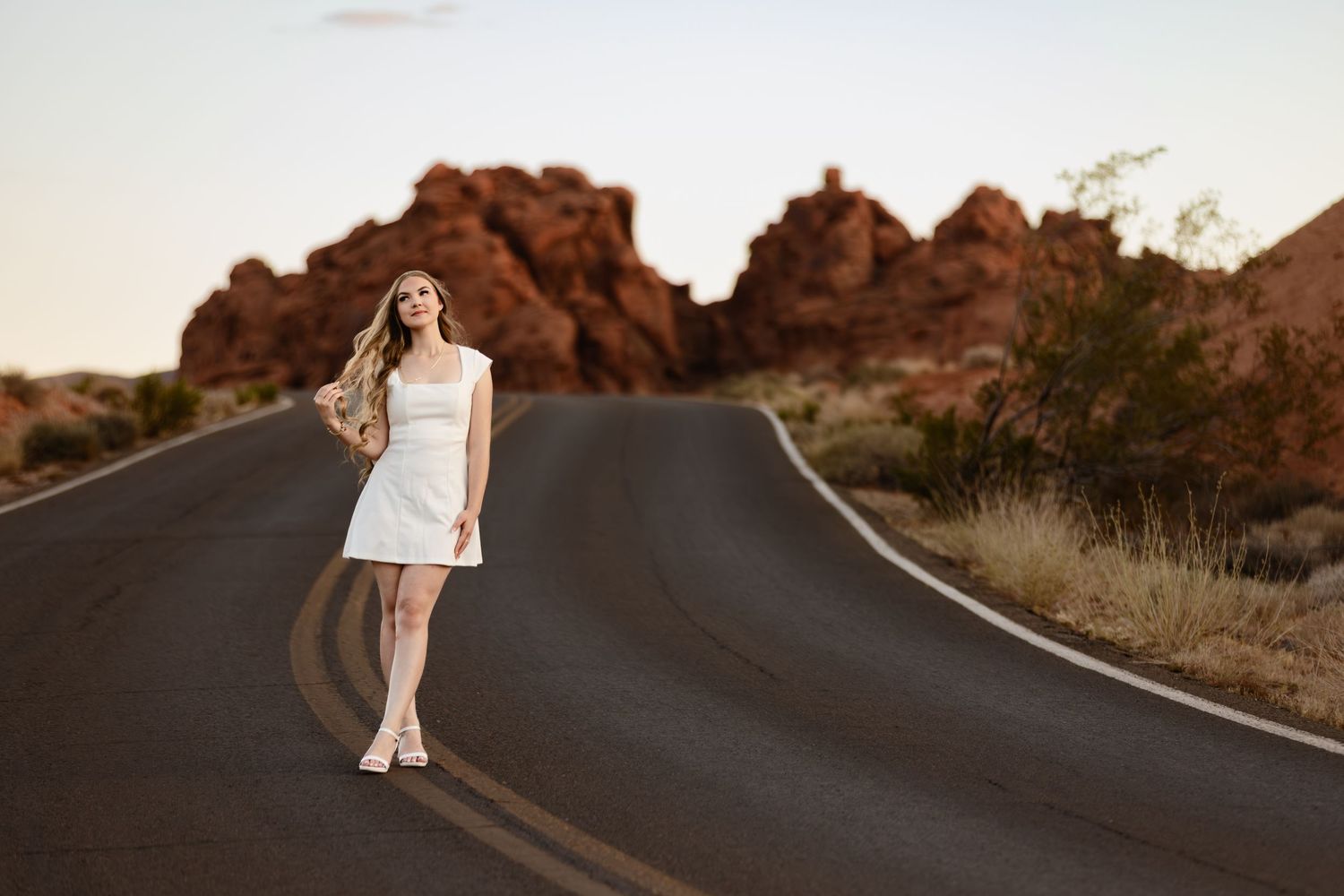 Young woman in white dress standing on winding desert road with red rock formations at sunset.