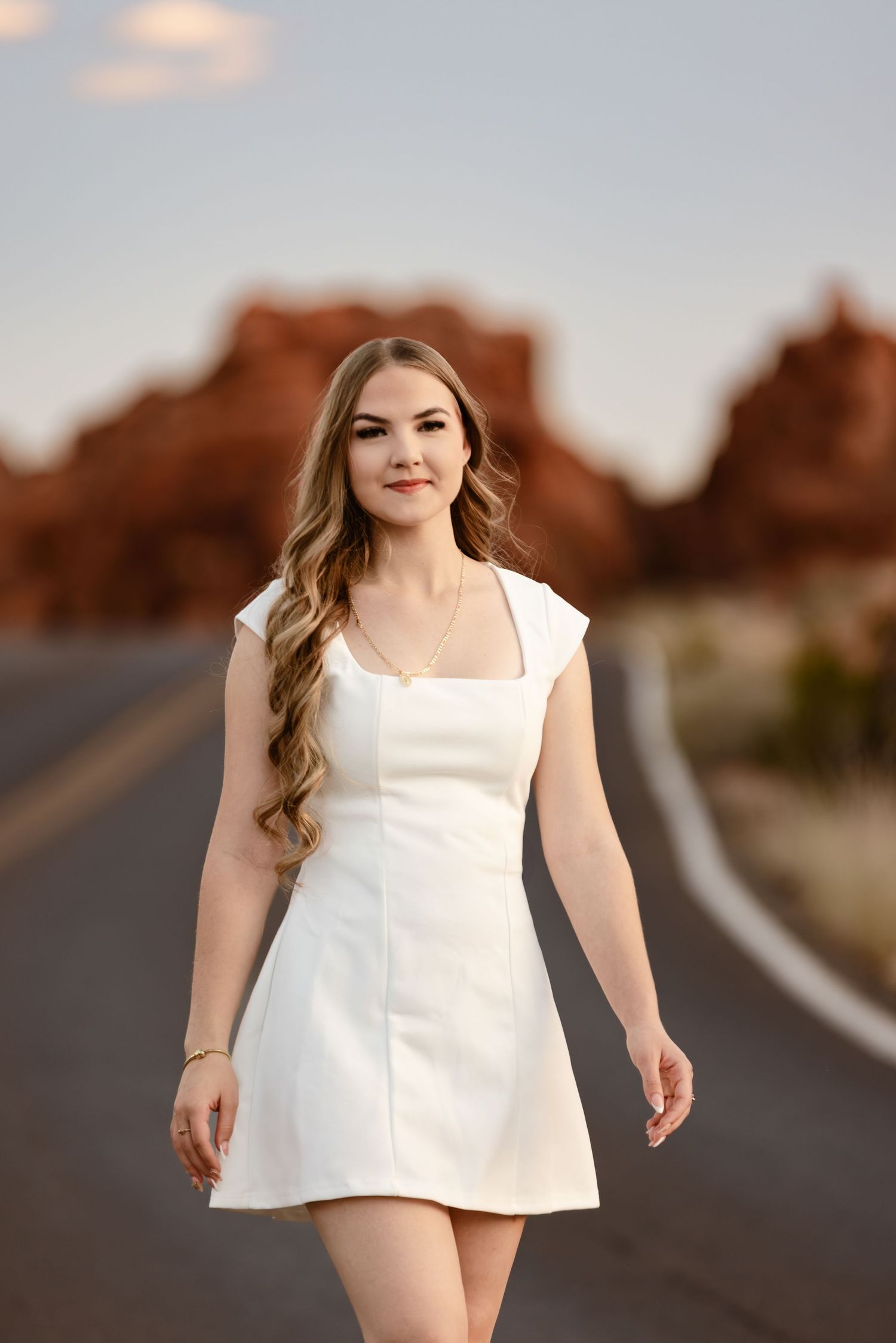 Young woman in white mini dress walking on desert road with red rock formations in background.