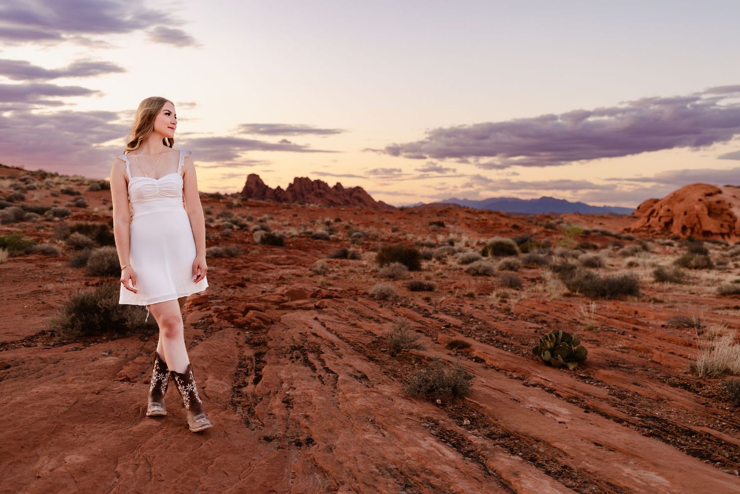 Woman in white dress and cowboy boots stands on red rock desert landscape at sunset with dramatic cloudy sky.