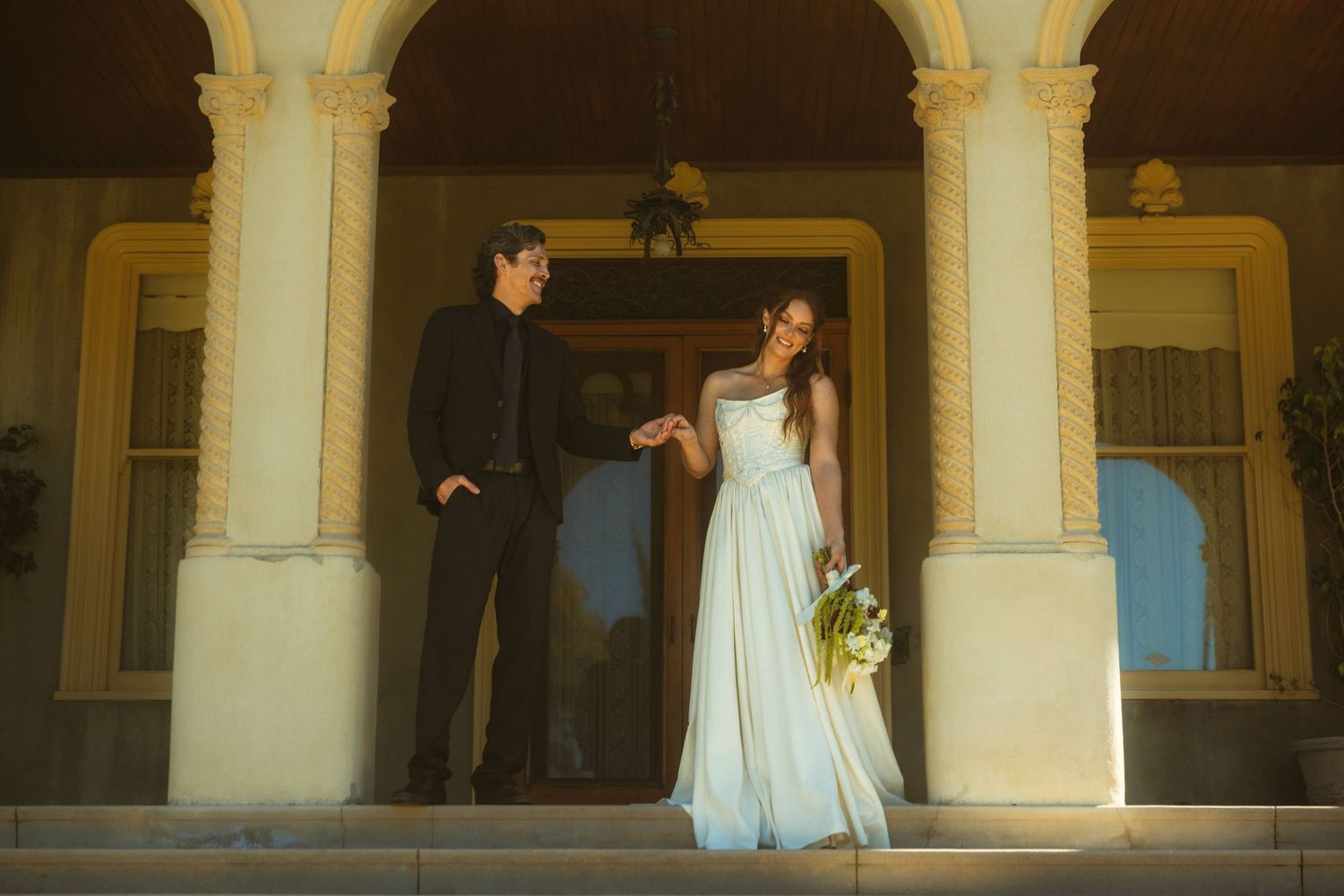 Elegant couple in wedding attire posed among sunlit columns, bride in white dress seated as groom stands behind her.
