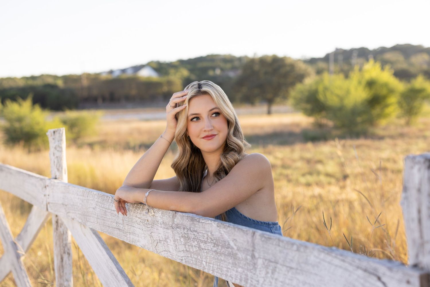 Young blonde woman leaning on white wooden fence in golden field during sunset portrait photography.