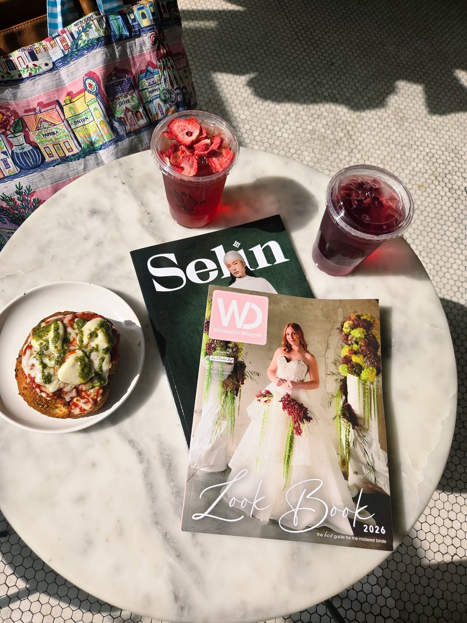 Marble table with Selin and WD Look Book 2026 magazines, a pizza toast, and two red berry drinks in clear cups.