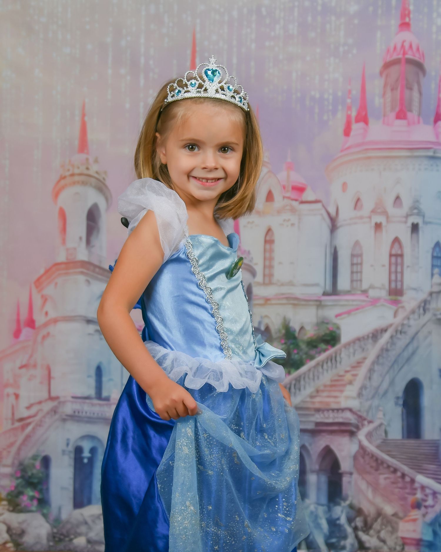 Young girl in blue Cinderella dress and tiara poses in front of fairy tale castle backdrop for princess photo shoot.