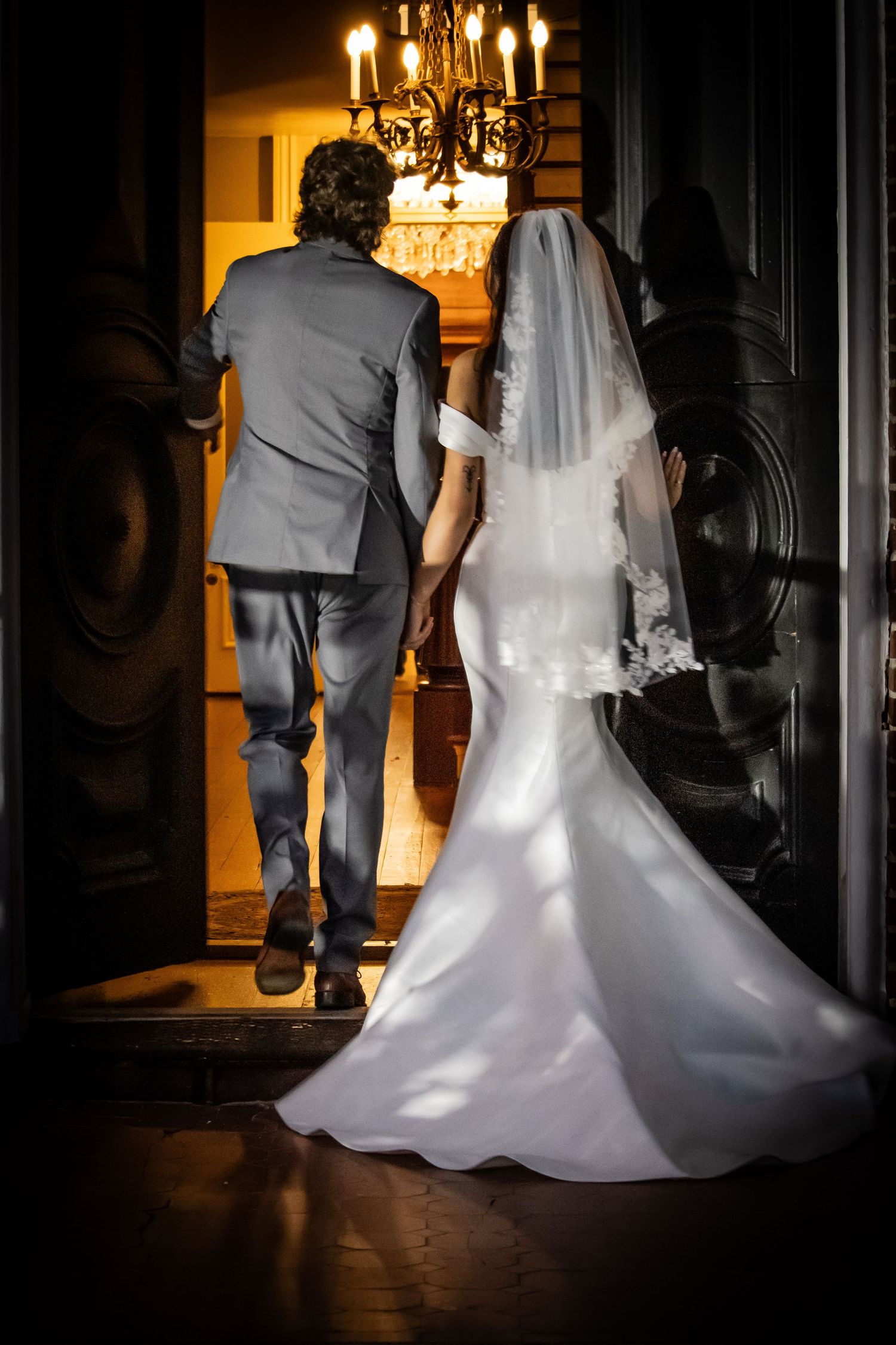 Bride in white gown and veil walks with groom in gray suit through ornate doors under chandelier.