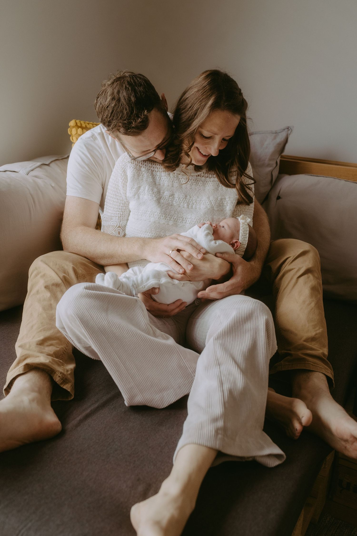 New parents sitting on a couch together, lovingly cradling their newborn baby in a warm, intimate home setting.