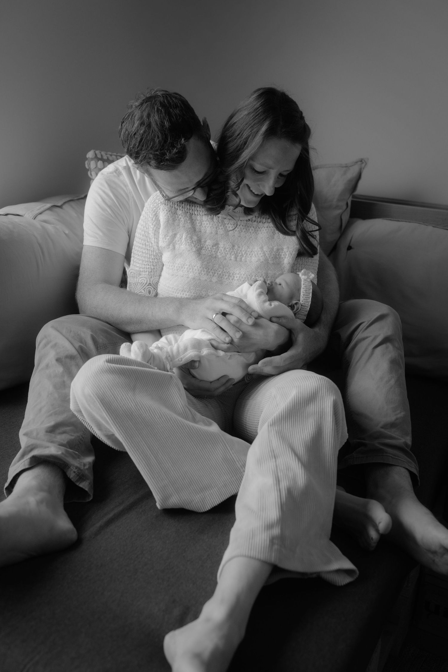 Black and white photo of parents sitting on couch lovingly gazing at their newborn baby.