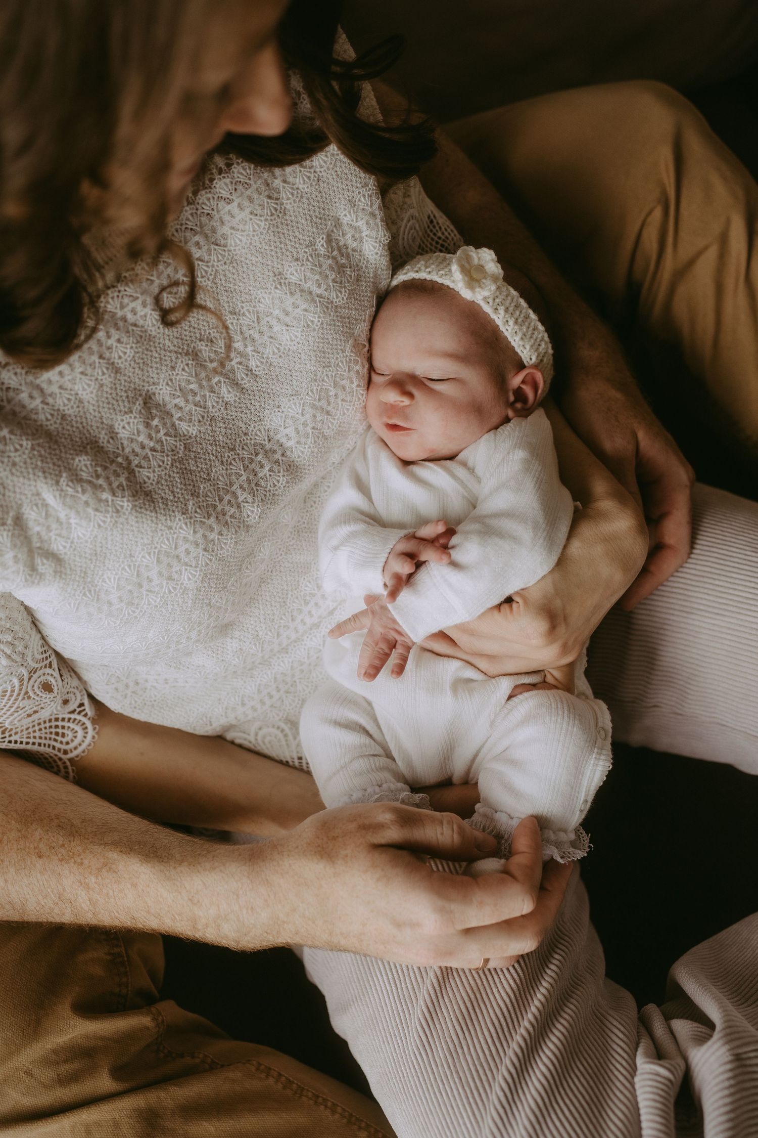 Newborn baby in white outfit and headband sleeping peacefully in parents arms during a warm lifestyle photoshoot.