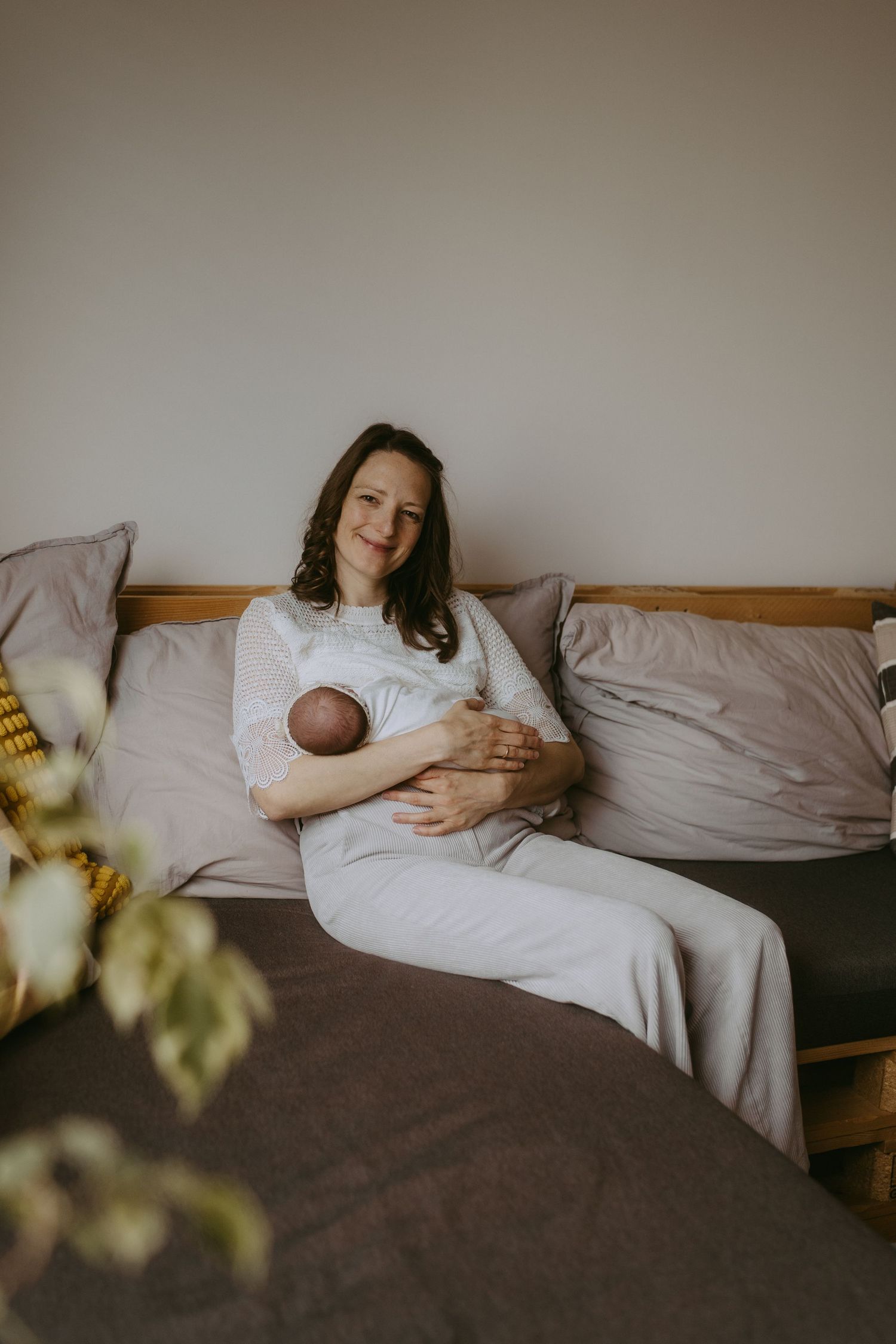 A smiling mother in a white dress sits on a bed cradling her newborn baby, in a cozy, softly lit bedroom.