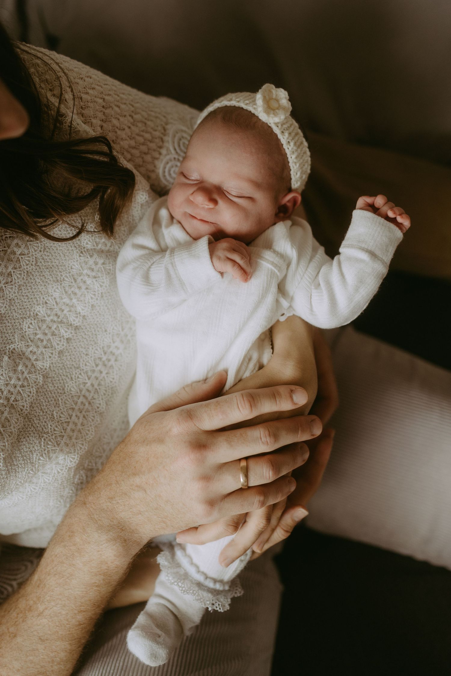 Newborn baby in white outfit and knit bonnet sleeping peacefully in parents hands, one wearing a gold wedding ring.
