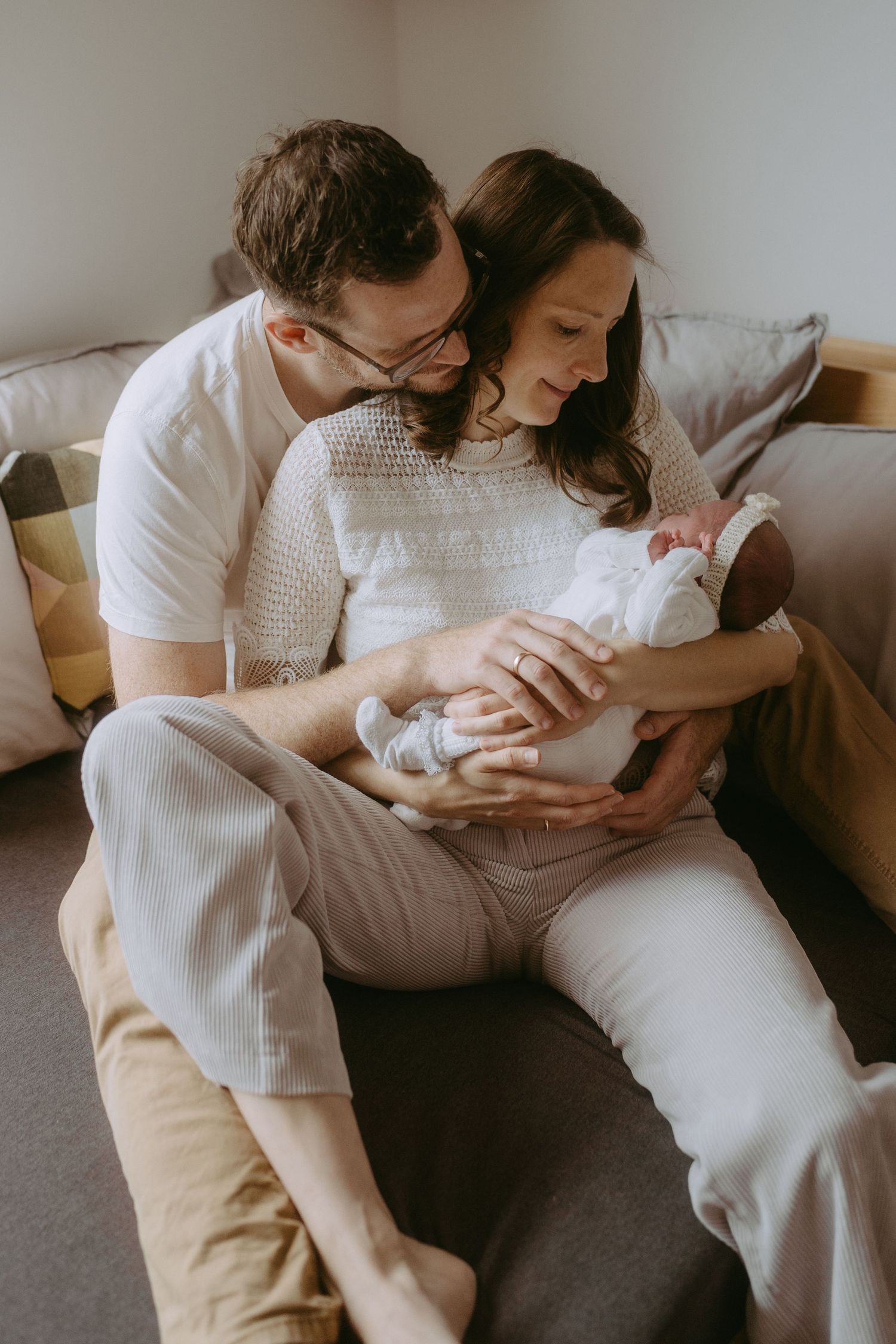 New parents sitting on a couch together, lovingly gazing at their newborn baby wrapped in white.