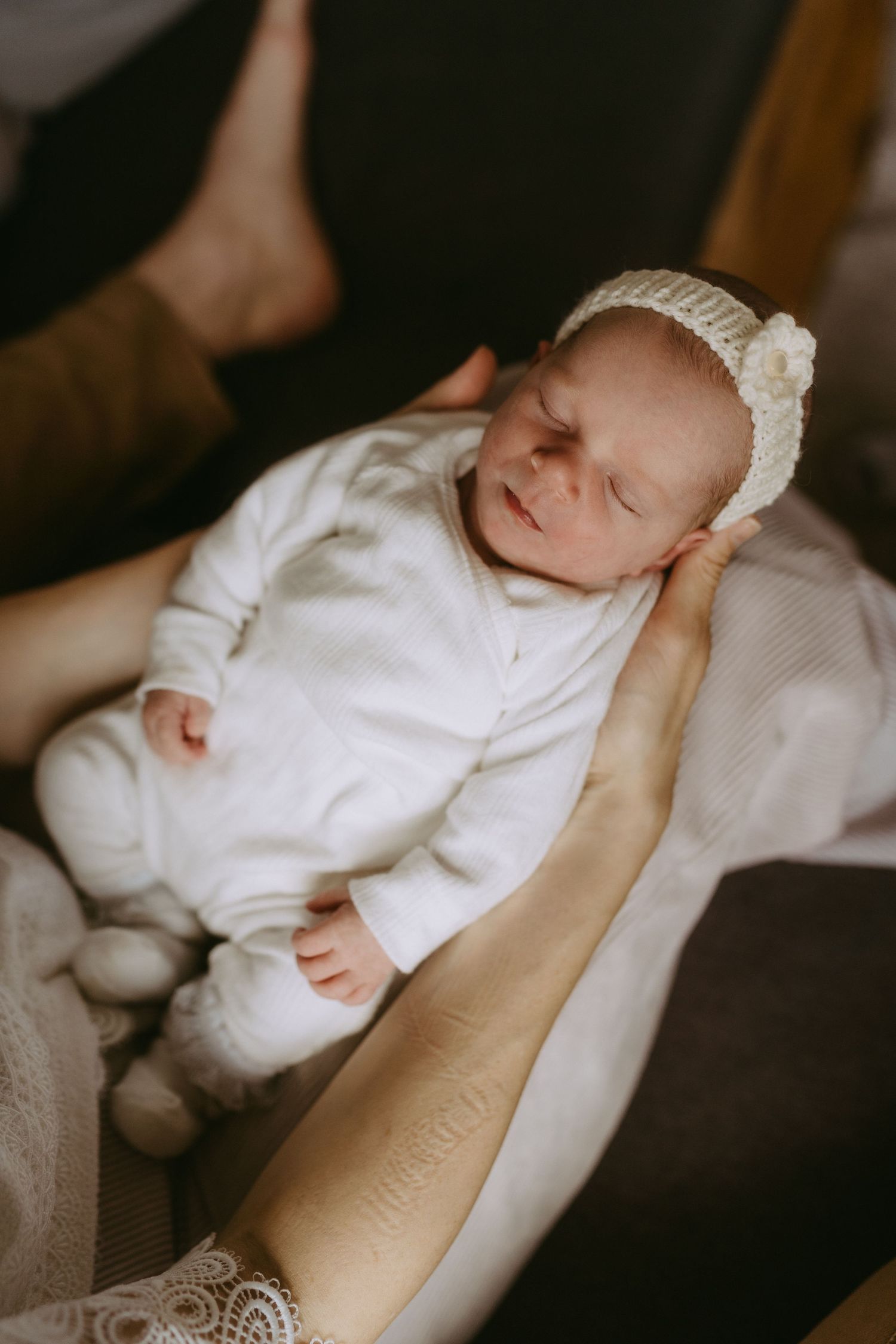 Newborn baby sleeping peacefully in white onesie and knit bonnet, cradled in parent's arms.