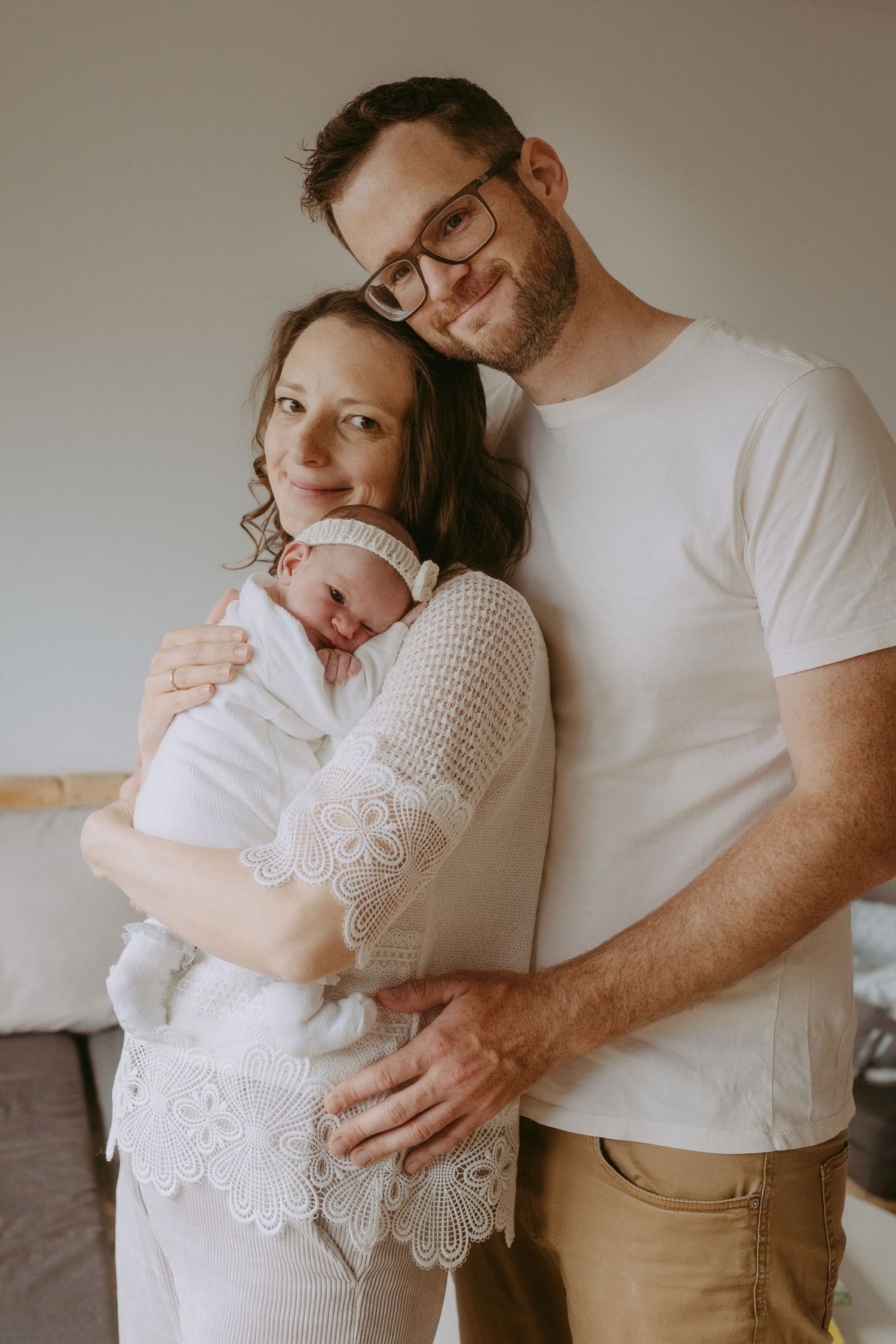 Happy couple in white outfits smiling while cradling their sleeping newborn baby at home.
