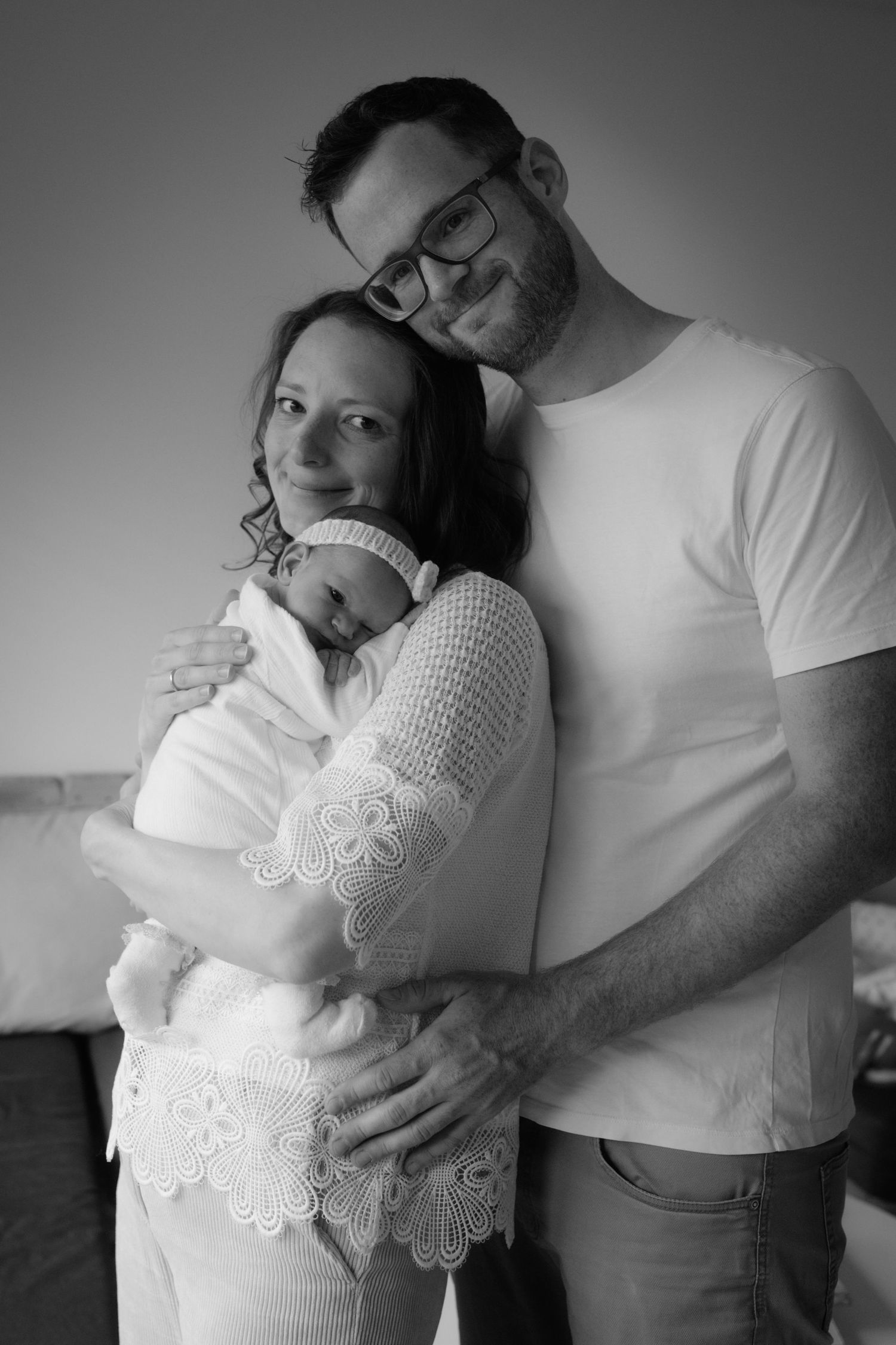 Black and white photo of new parents smiling and holding their swaddled newborn baby at home.
