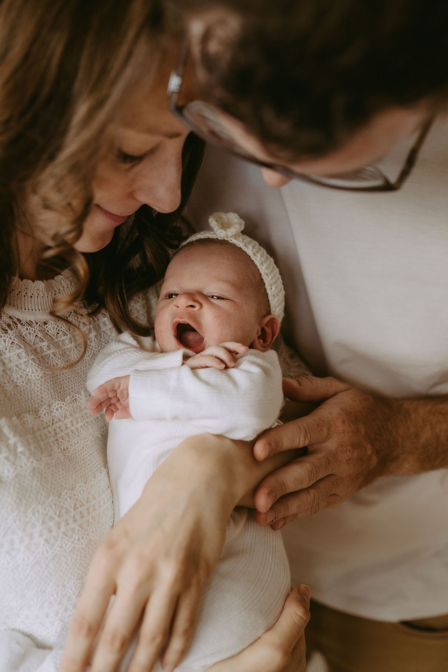 Parents lovingly gaze at their yawning newborn baby dressed in white, sharing a tender family moment.