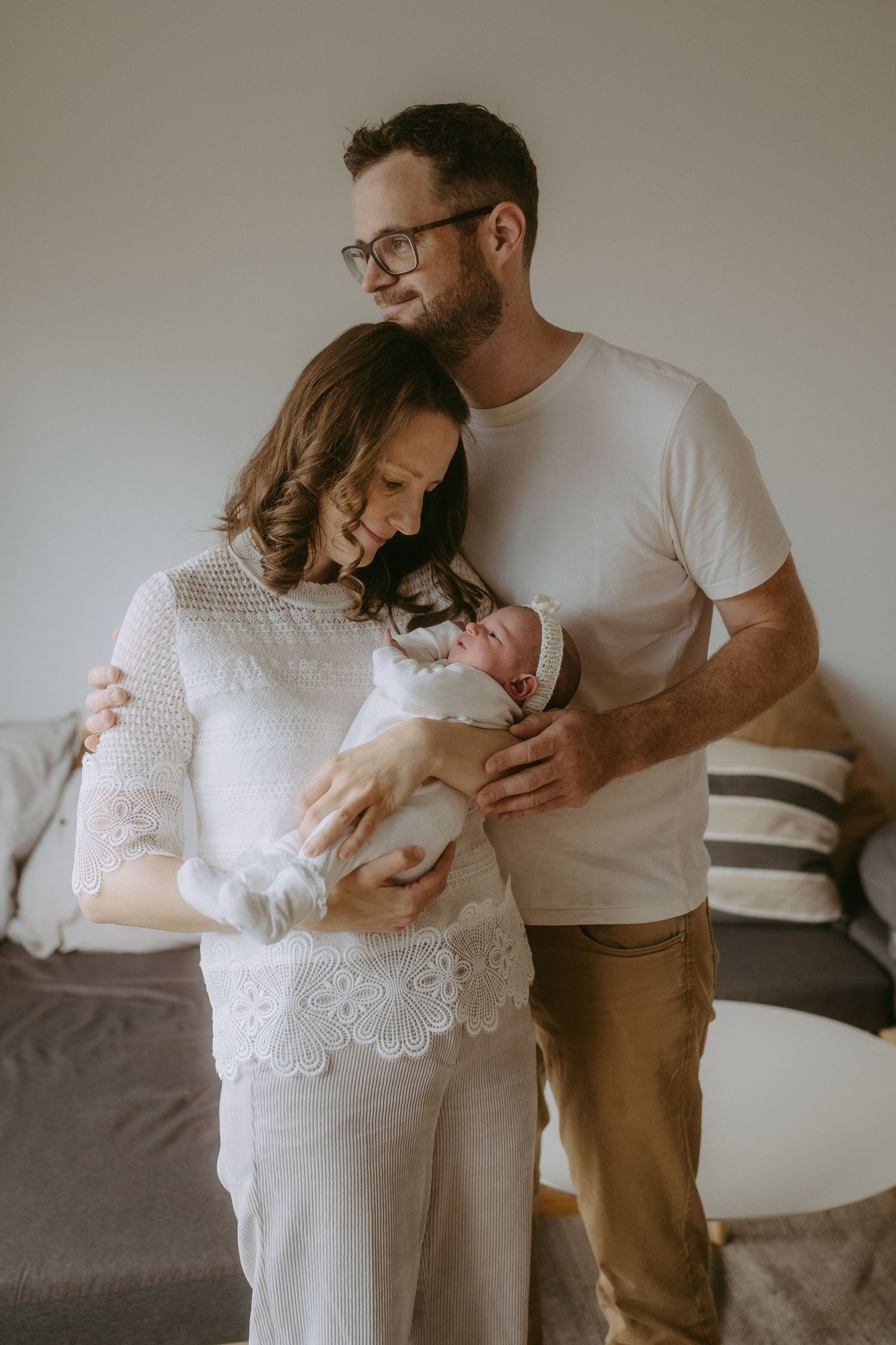 New parents holding their swaddled newborn baby together, mother in white lace dress, father in white t-shirt.