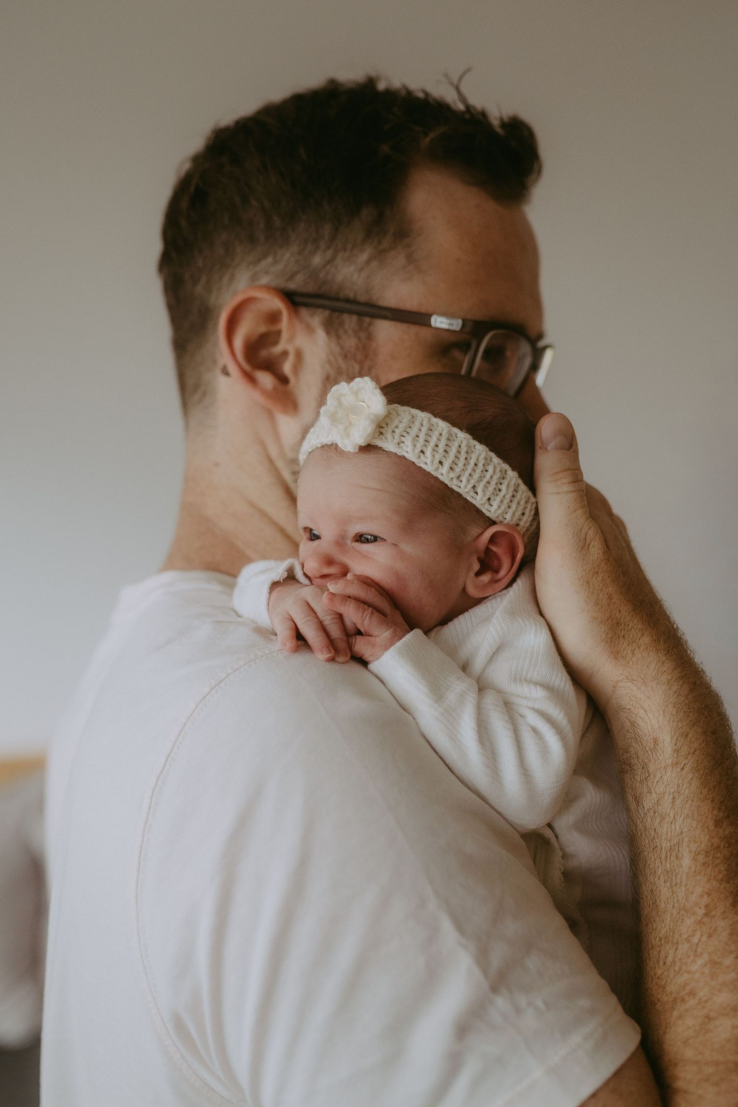 A father in a white t-shirt gently holds his newborn baby girl with a floral headband against his shoulder.