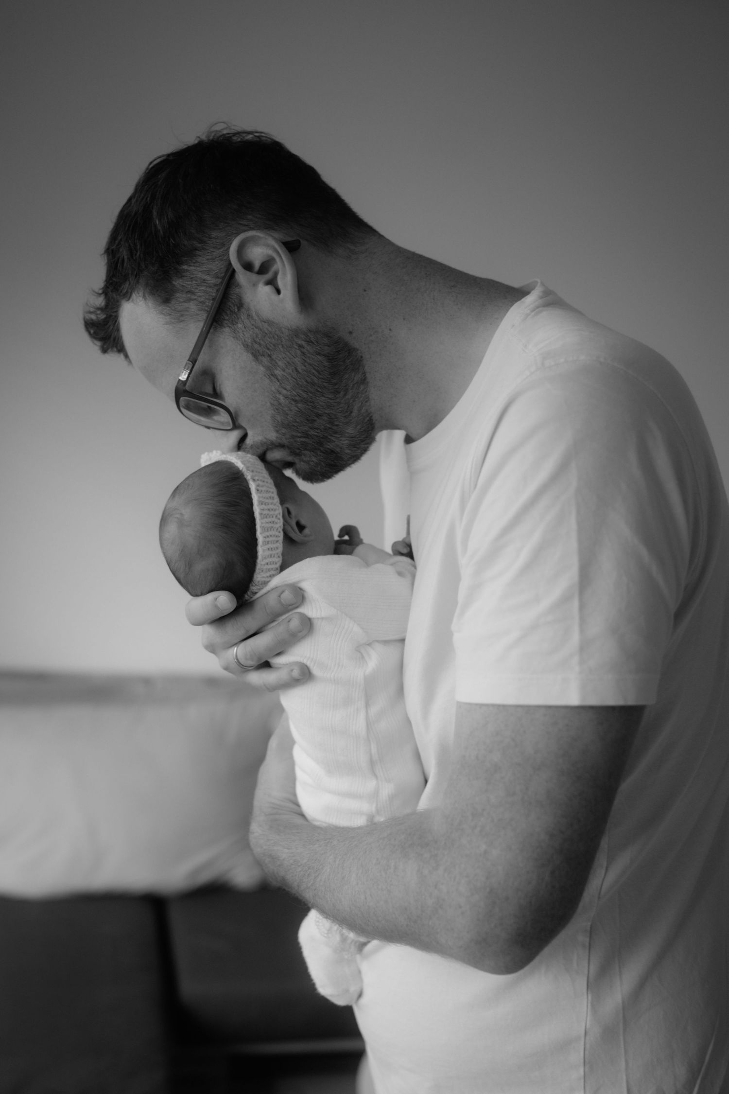 Black and white photo of a bearded father tenderly kissing his newborn baby while cradling it gently in his arms.