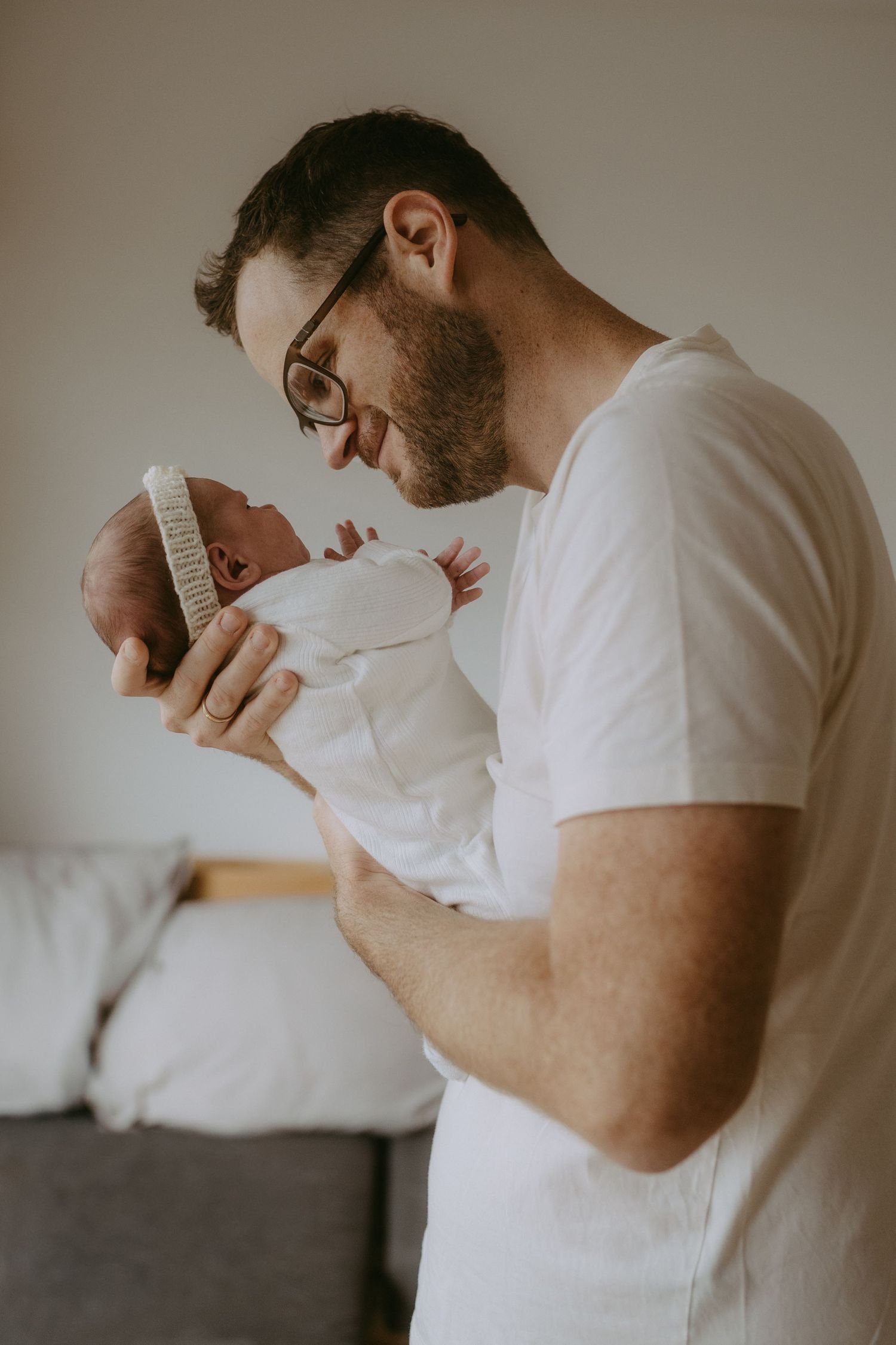 New father in white t-shirt and glasses tenderly holding his swaddled newborn baby close to his face.