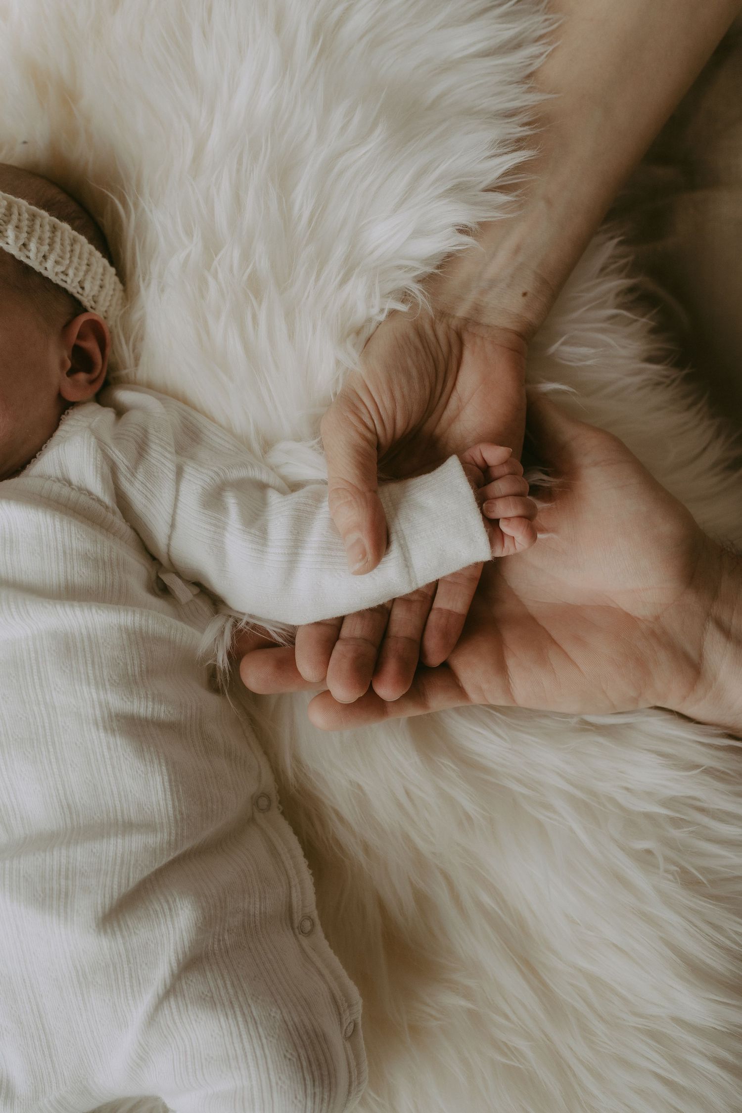 A newborn baby in white onesie lying on fluffy fur rug while adult hands gently hold its tiny feet.