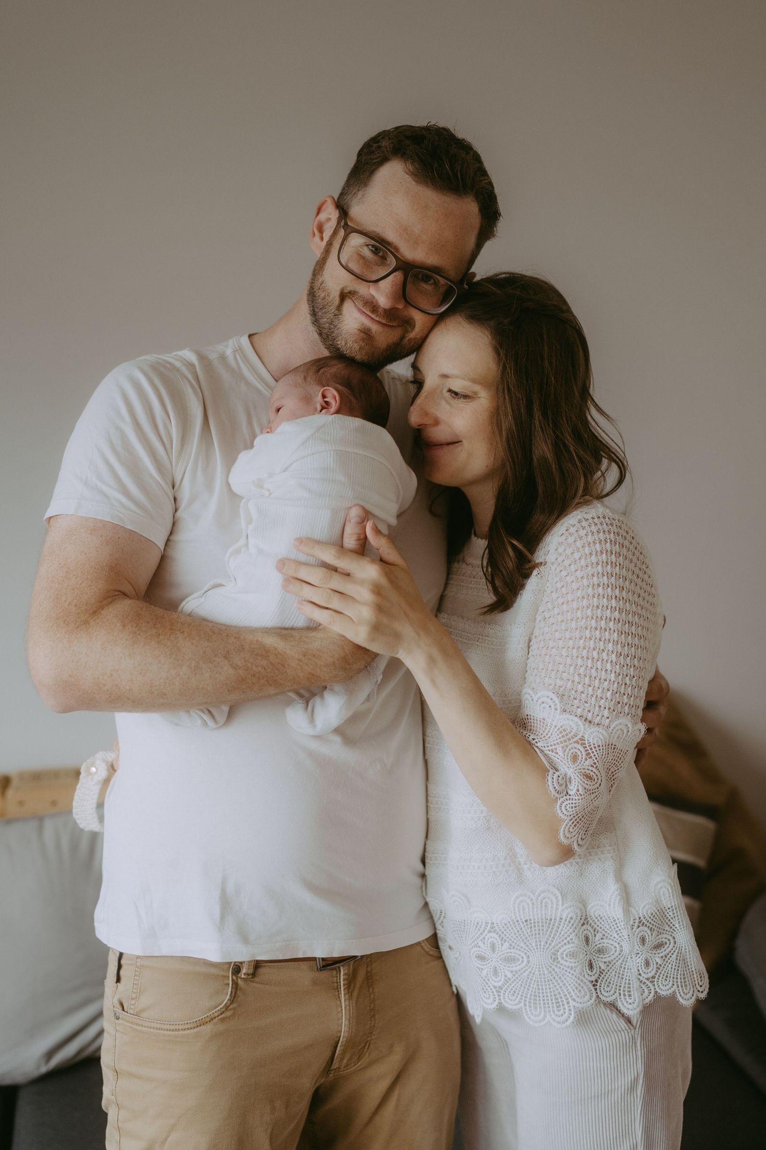 Happy couple in white outfits tenderly holding their newborn baby together in a bright indoor setting.