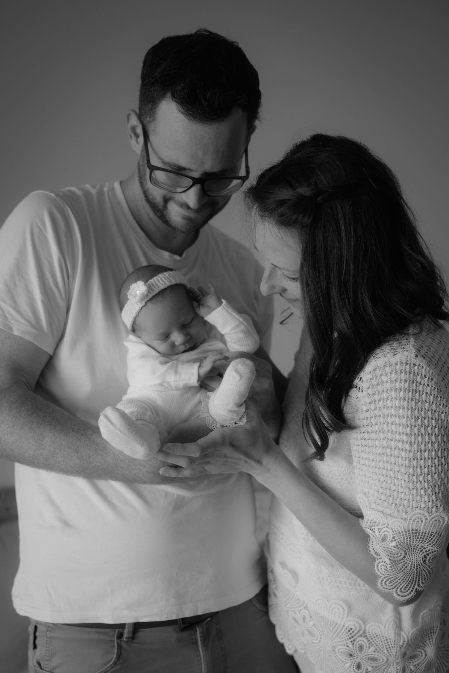 Black and white portrait of new parents smiling down at their sleeping newborn baby in fathers arms.