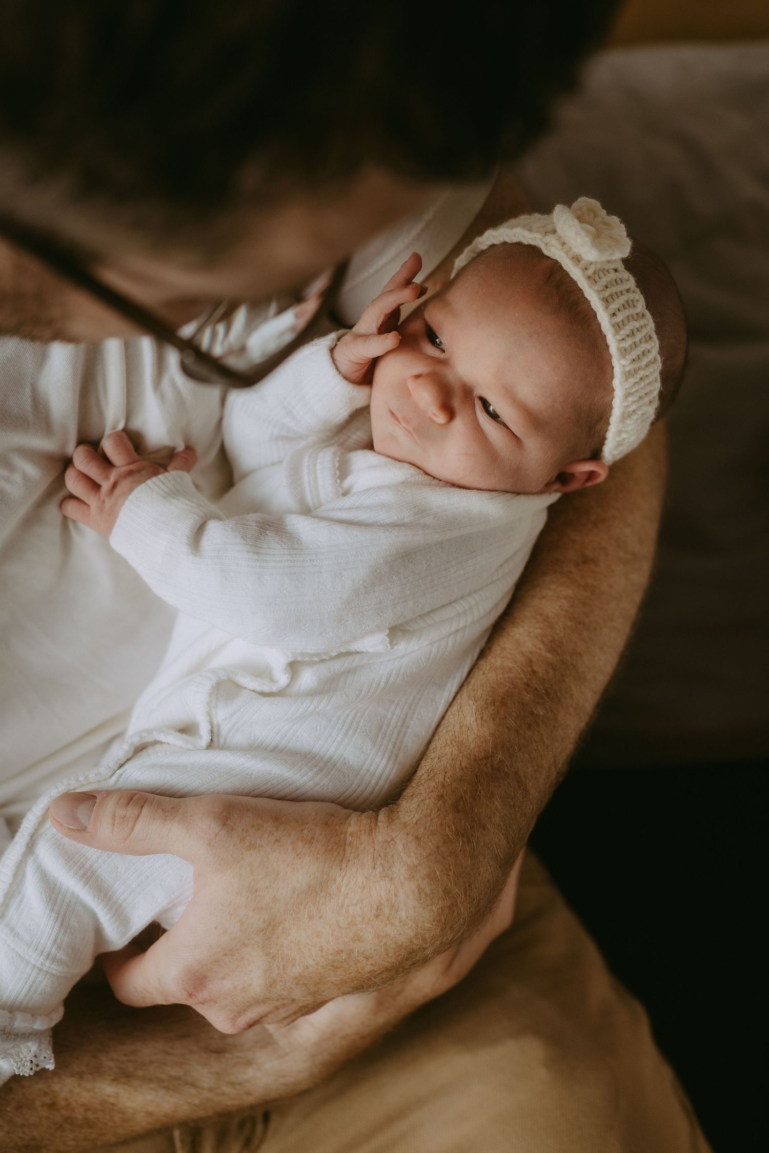 Newborn baby in white onesie and cream knit bonnet smiling while being cradled in a parent's arms.