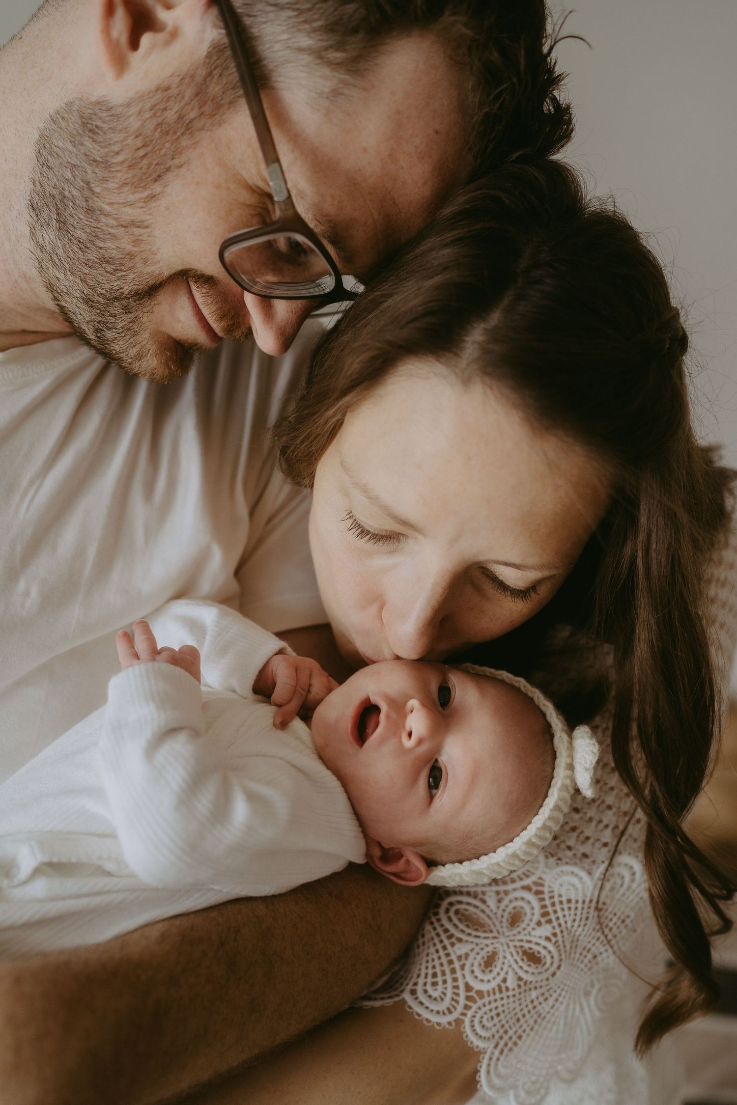 New parents lovingly gazing at and kissing their alert newborn baby wrapped in white cloth.