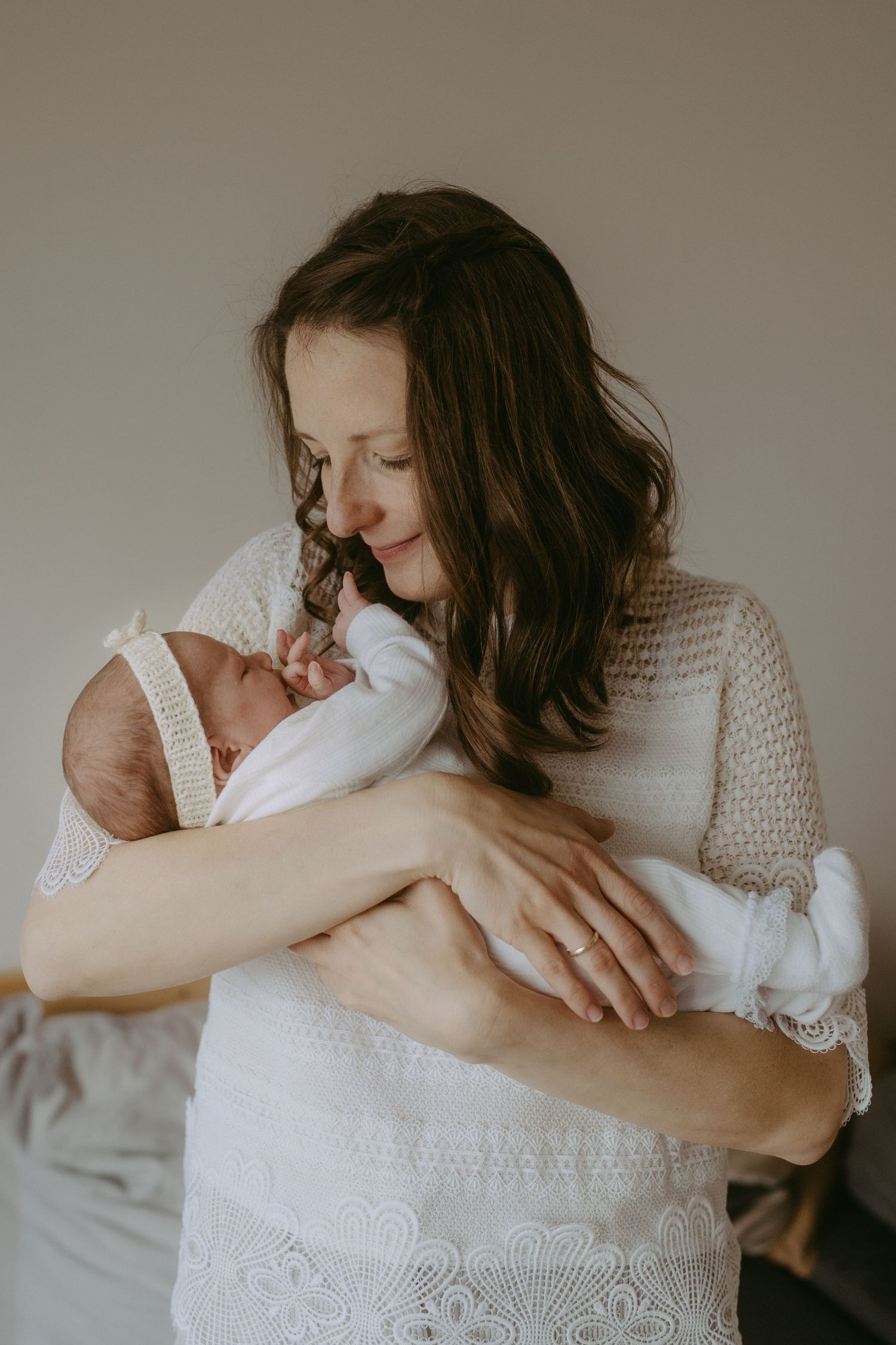 A mother in a white lace dress tenderly holds her swaddled newborn baby, gazing down with a warm smile.