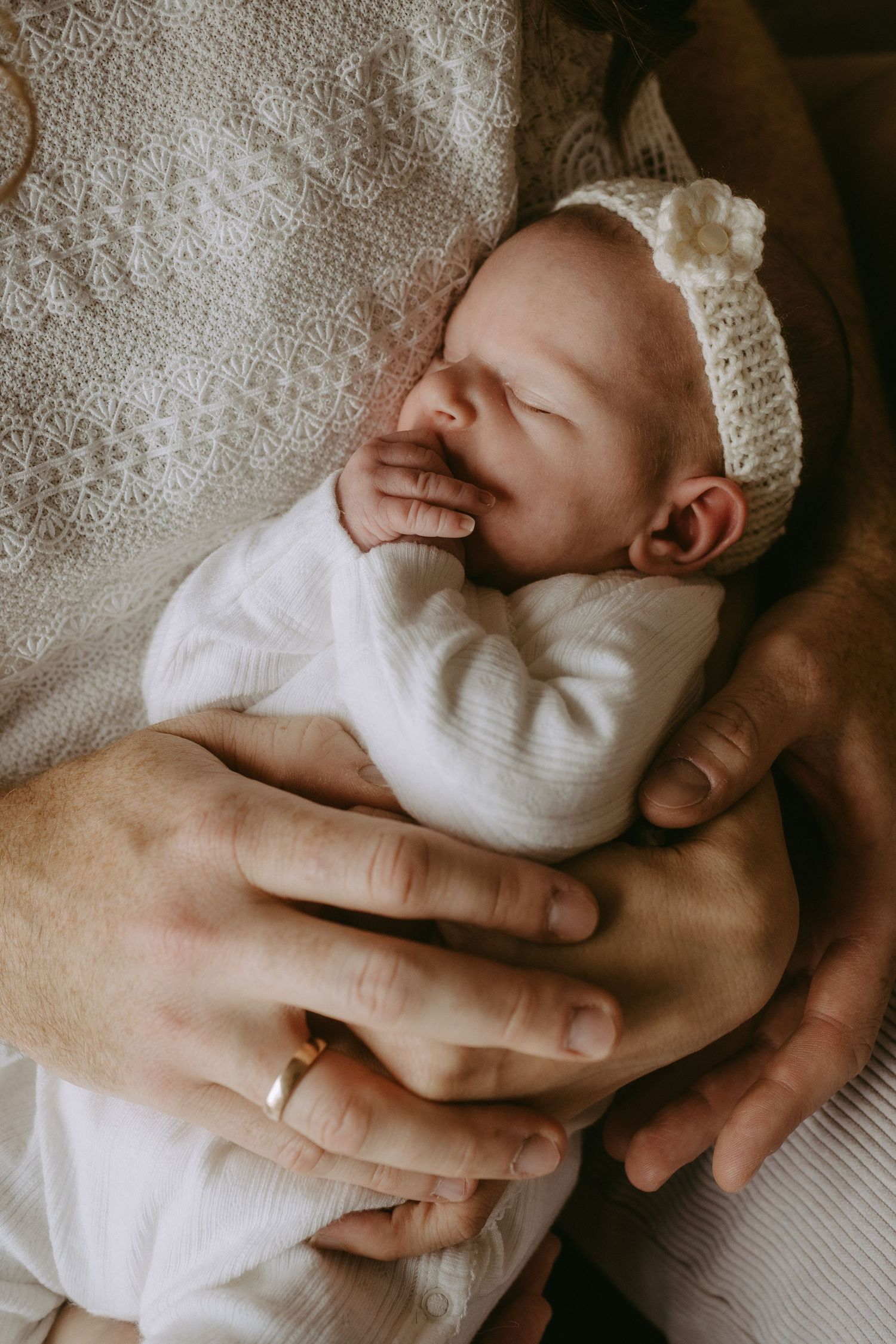 Newborn baby in white knit outfit and floral headband resting peacefully in parents hands.