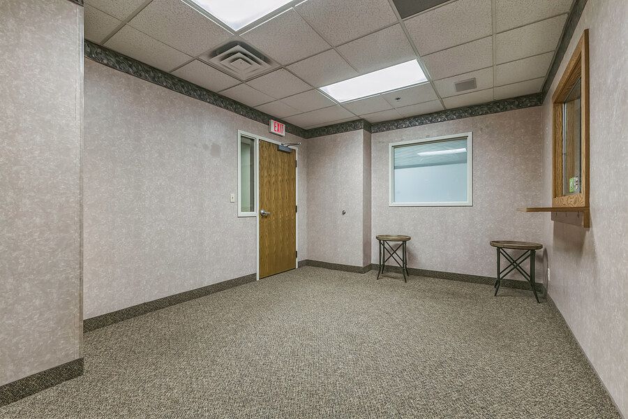 Empty commercial office room with speckled gray carpet, drop ceiling, wooden door, and small window in a neutral-toned space.