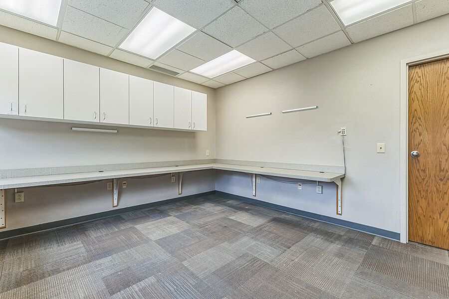 Spacious commercial office room with white upper cabinets, built-in countertops, drop ceiling with skylights, and carpeted flooring.