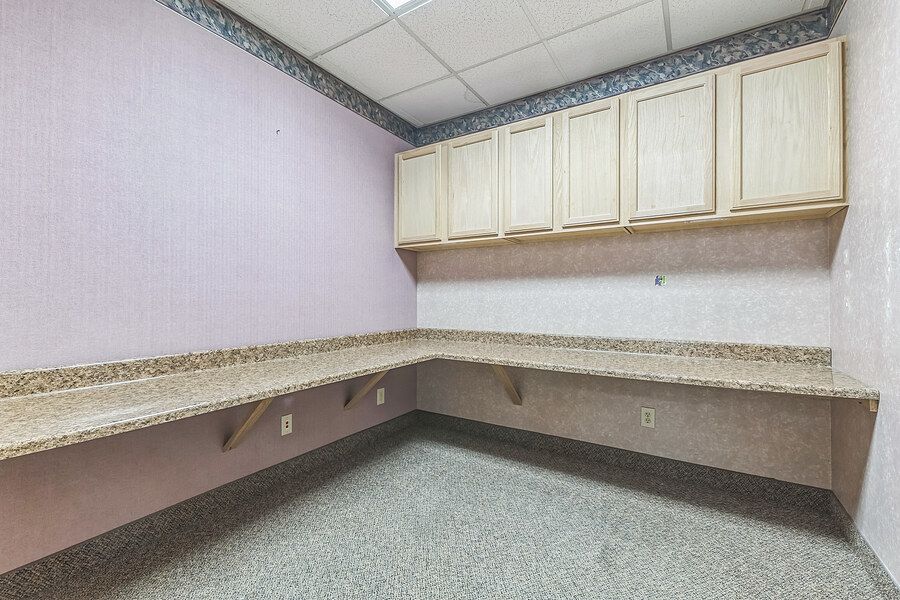 Corner office workspace with built-in granite countertop desk, light wood upper cabinets, and neutral carpet flooring.