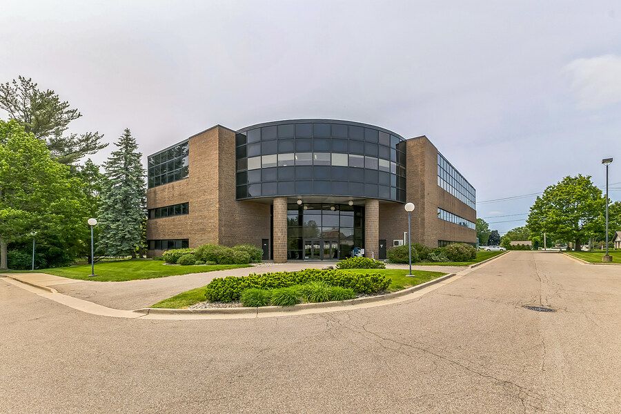 Modern brick office building with curved glass facade surrounded by lush green trees and landscaped grounds.